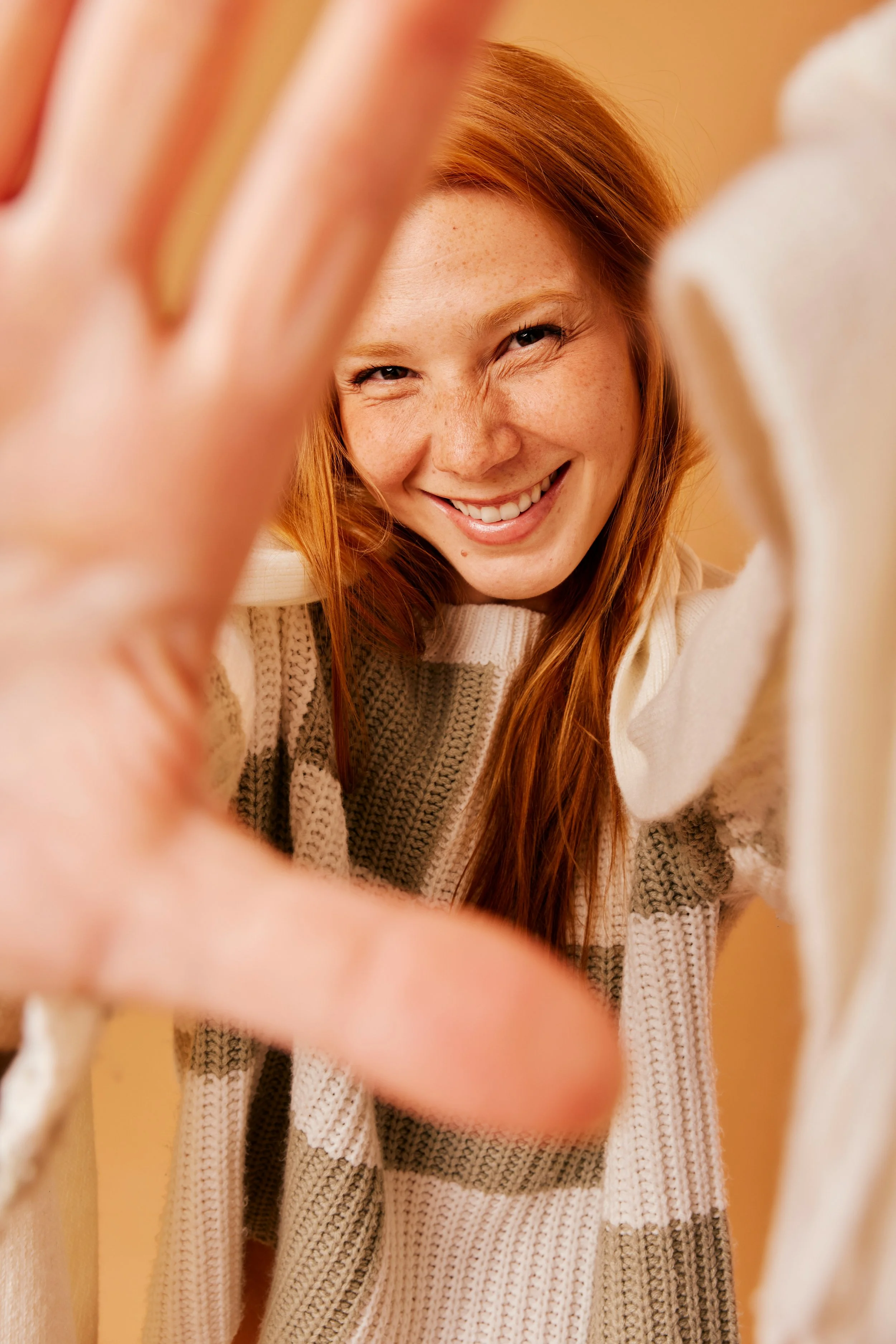 A young woman with red hair smiling and reaching towards the camera, wearing a striped knit sweater.