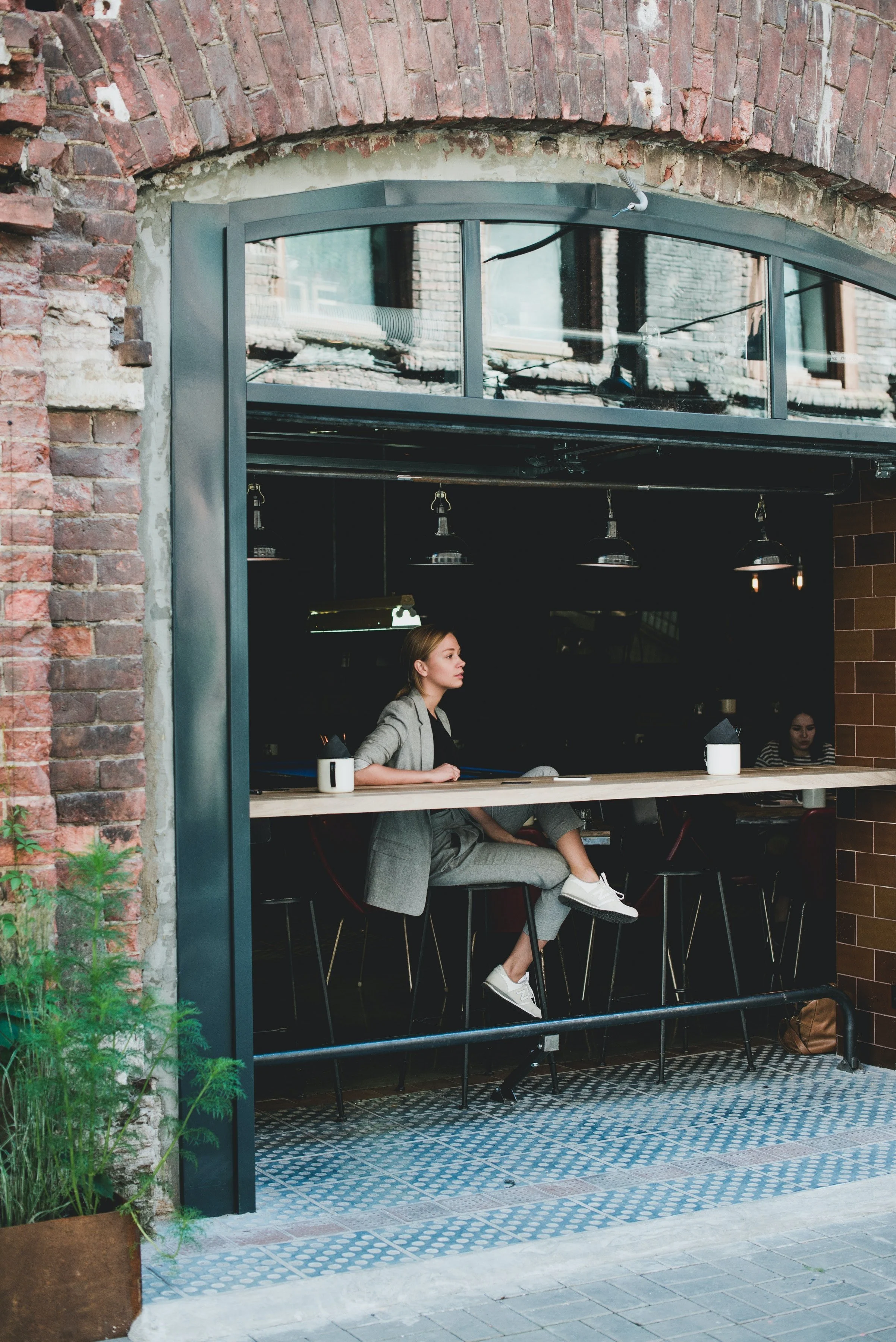 A woman sitting at a counter inside a brick-walled cafe, wearing a gray blazer, matching pants, and white sneakers, with a clear view of her profile and a group of people in the background.