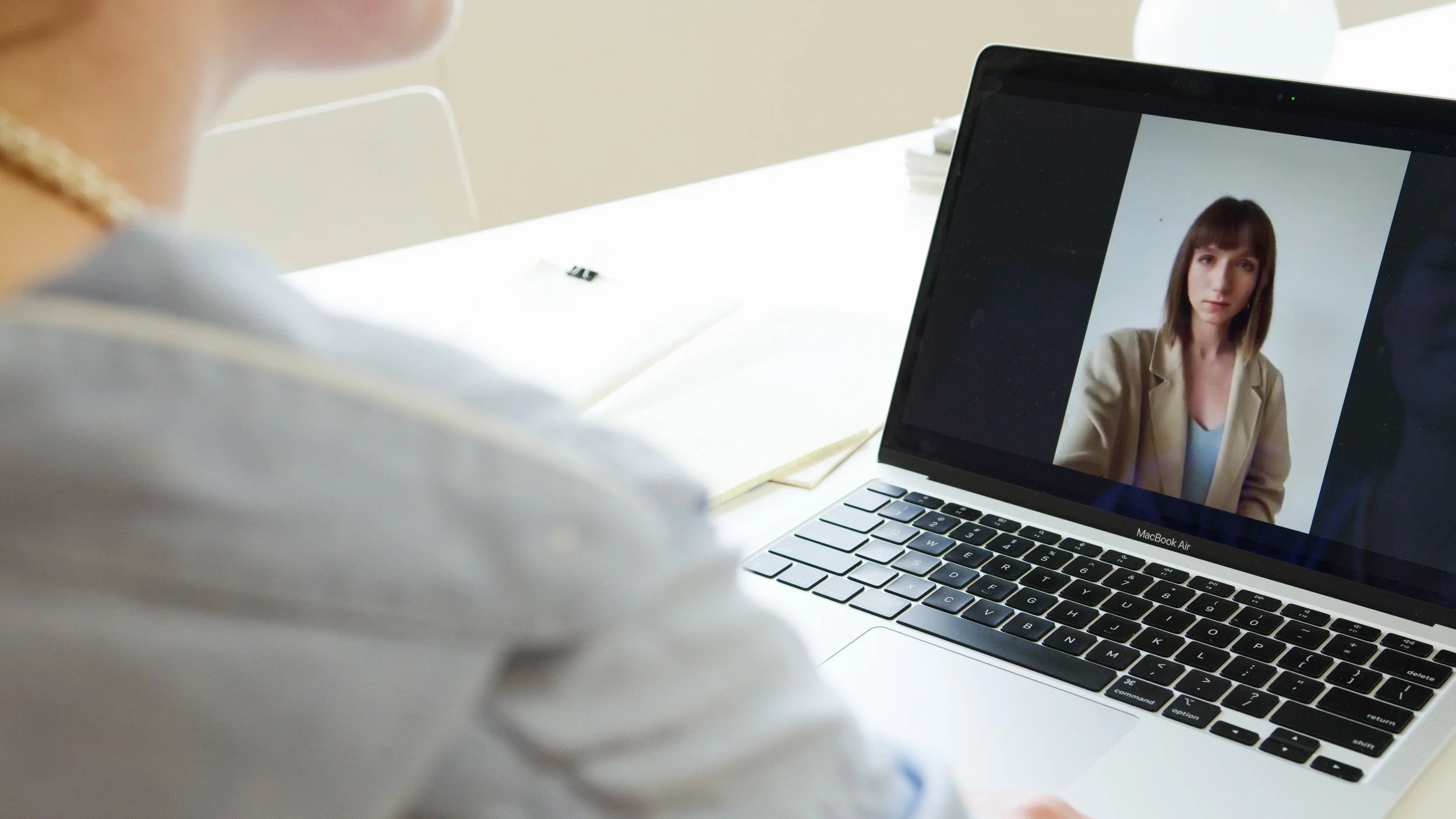 A person on a video call with a woman shown on a laptop screen, sitting at a white desk with notebooks.