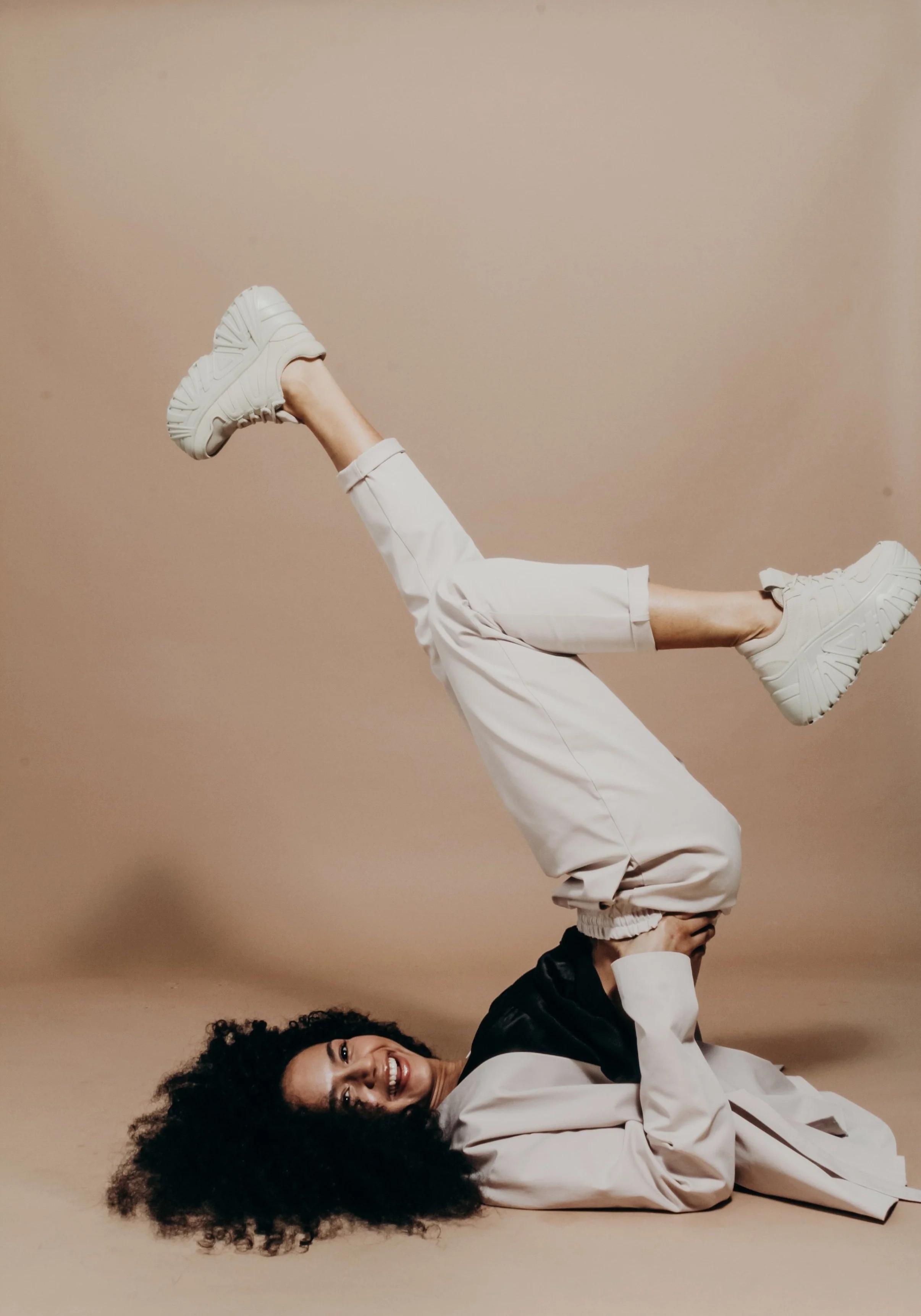 Woman with curly hair lying on the ground, smiling, in a pose with one leg raised and bent at the knee, wearing white sneakers, light-colored pants, and a black and beige jacket, against a beige background.