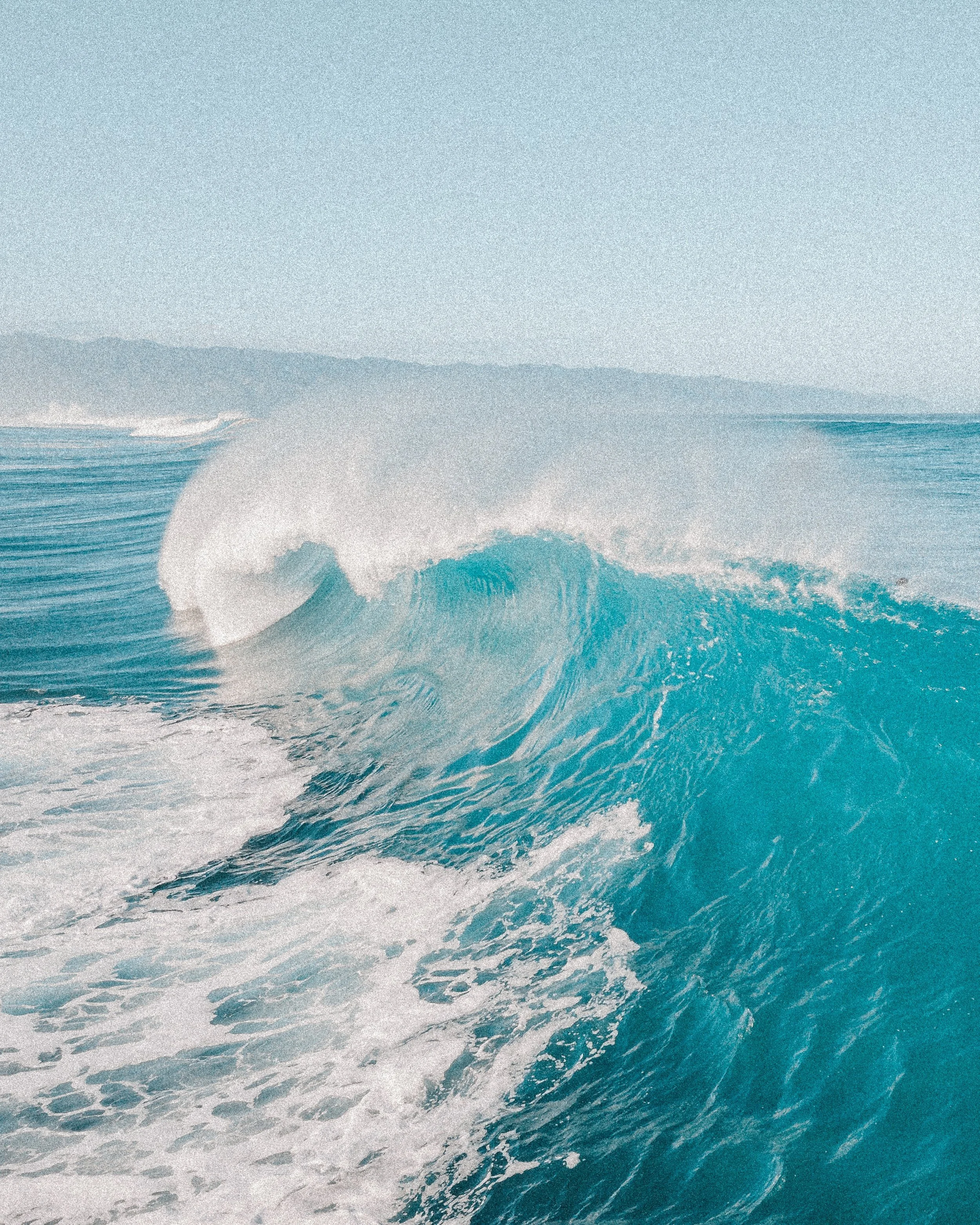 Ocean wave with white foam and a distant shoreline and mountains in the background.