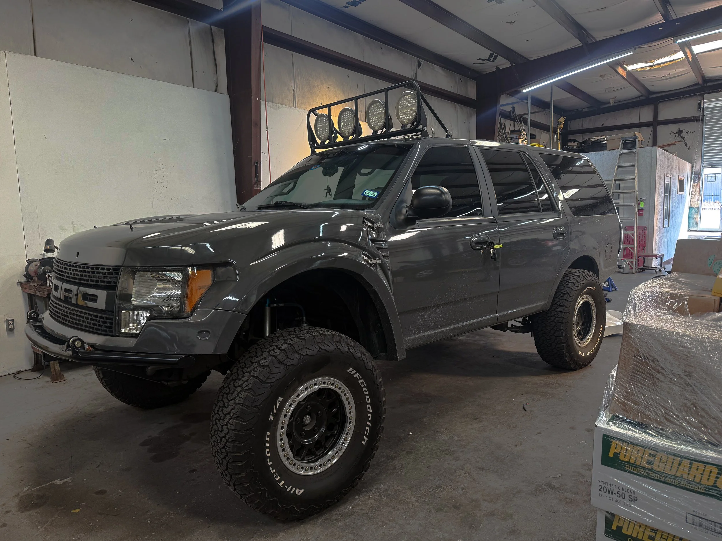 A dark gray Ford SUV with off-road tires and roof-mounted lights inside a garage, with various tools and supplies around.