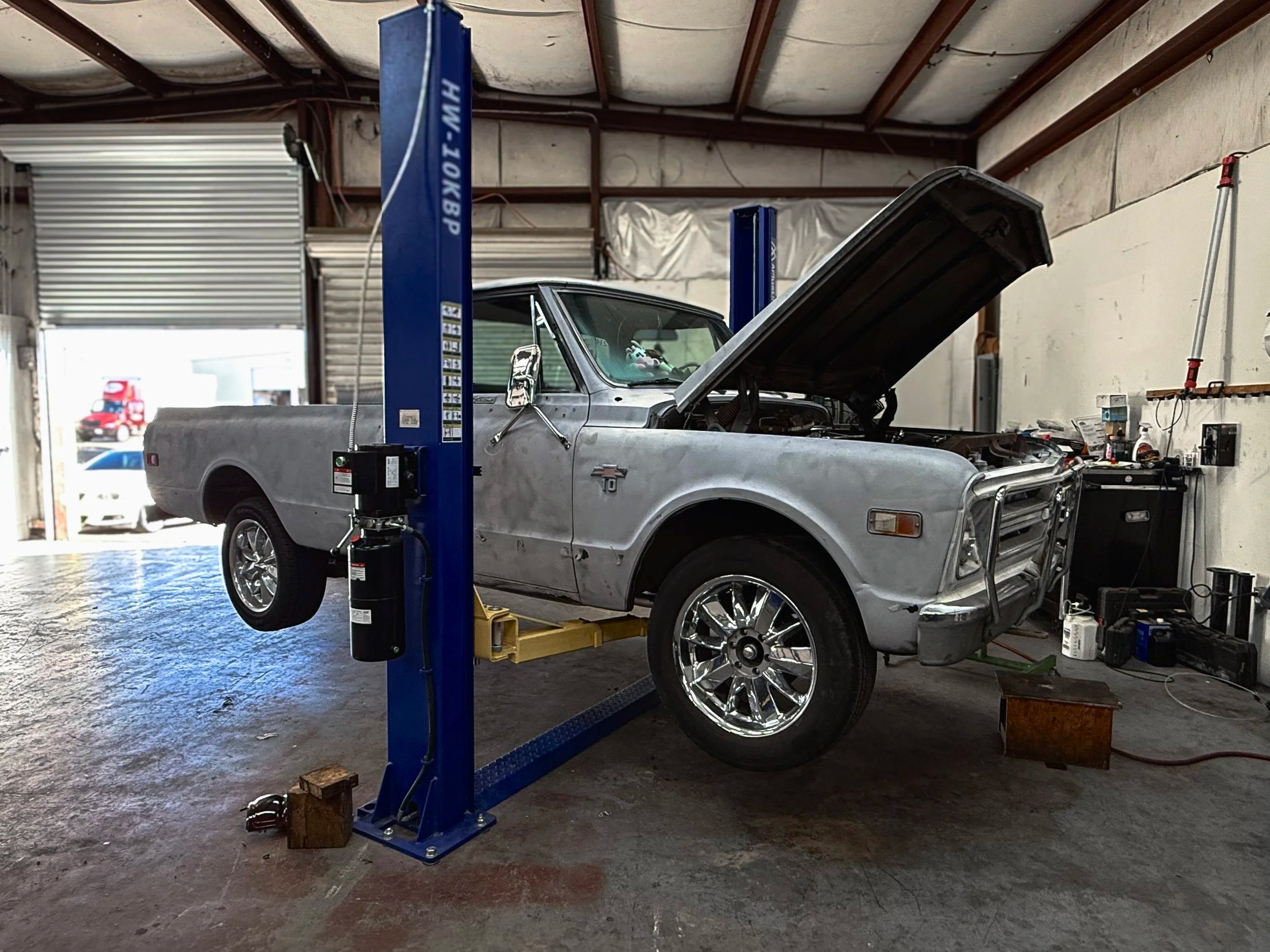 A vintage pickup truck with a weathered, unfinished body on a hydraulic lift in an automotive workshop, with the hood open and various tools and equipment nearby.