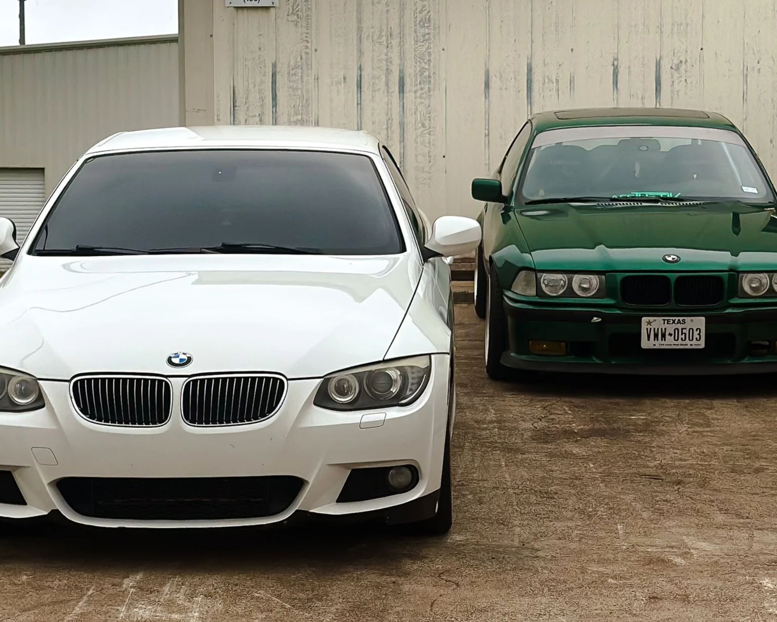 A white BMW 328i parked next to a green BMW 325is on a concrete surface, with a beige metal building in the background.