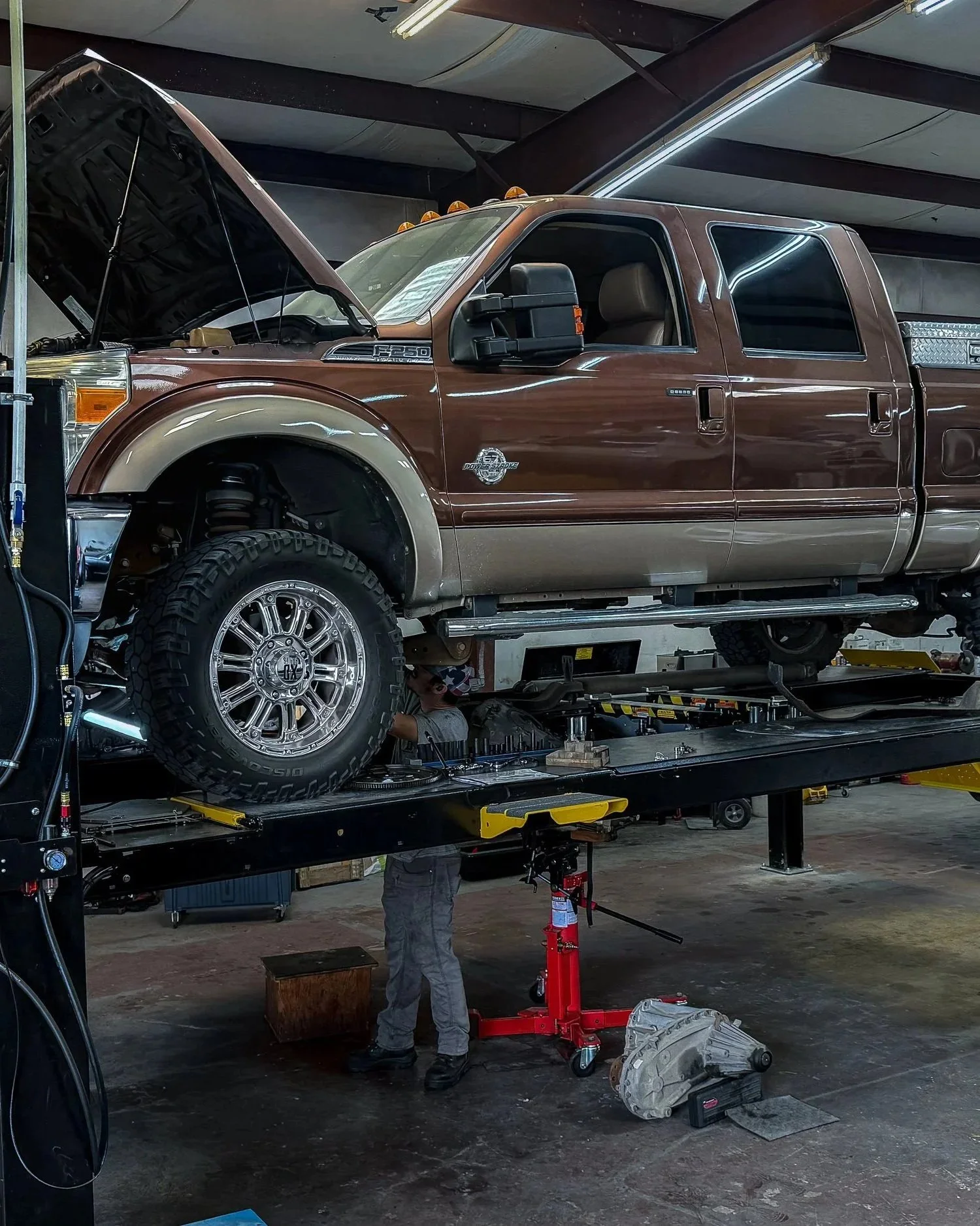 A mechanic working underneath a lifted brown and beige Ford F-250 truck in an auto repair shop.