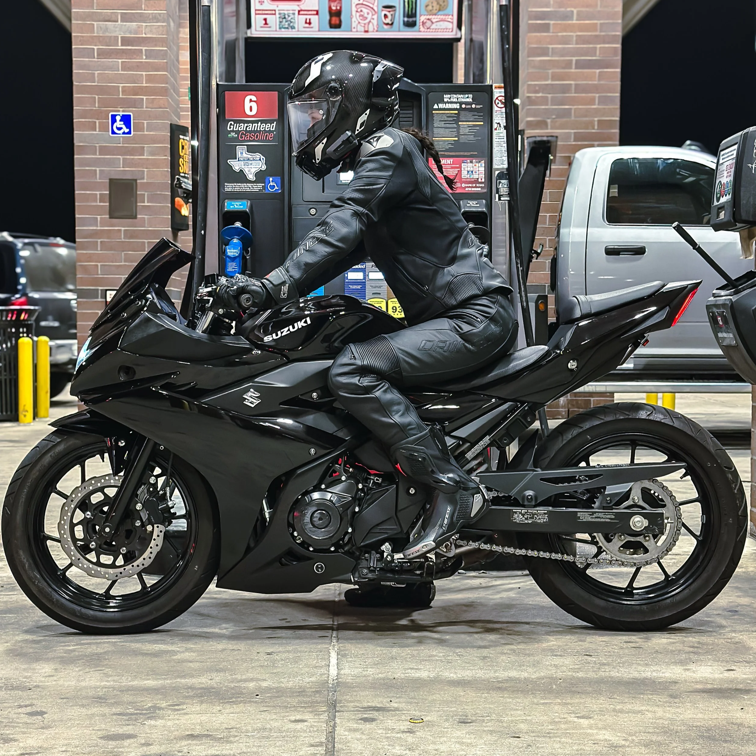 Motorcycle rider in full black riding gear sitting on a black Suzuki sportbike at a gas station
