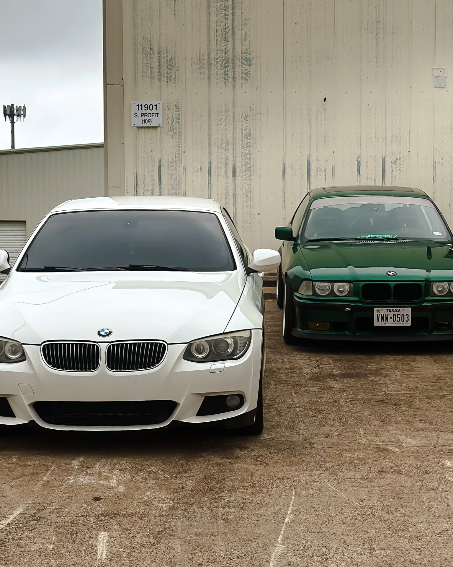 A white BMW 328i car parked next to a green BMW 325is car in an outdoor lot against a beige wall with a sign that reads '11901 S. Profit (109)'.