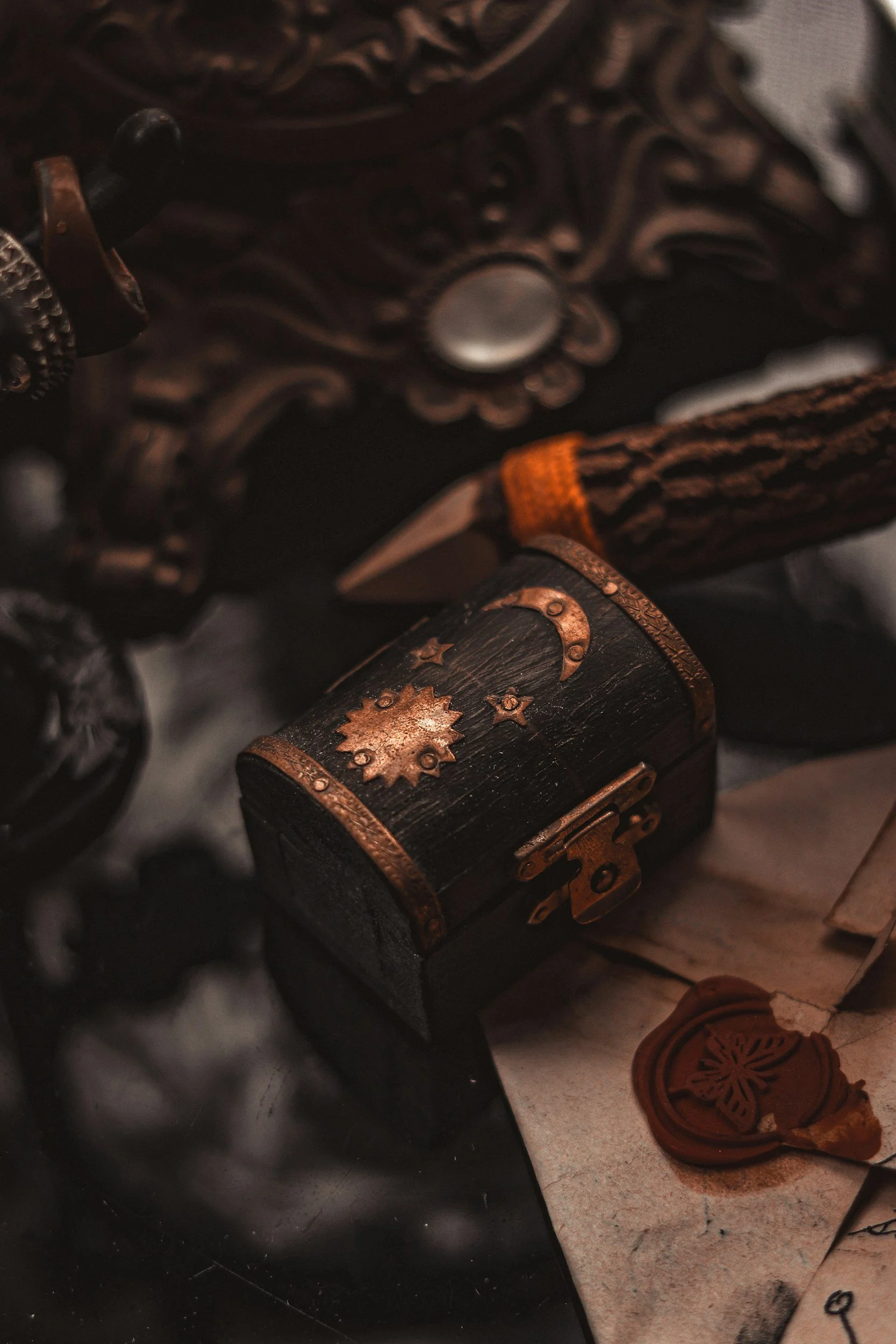 Small black chest with copper and gold decorative hardware, placed on aged paper, with a wax seal nearby. Part of ornate dark wood frame in background.