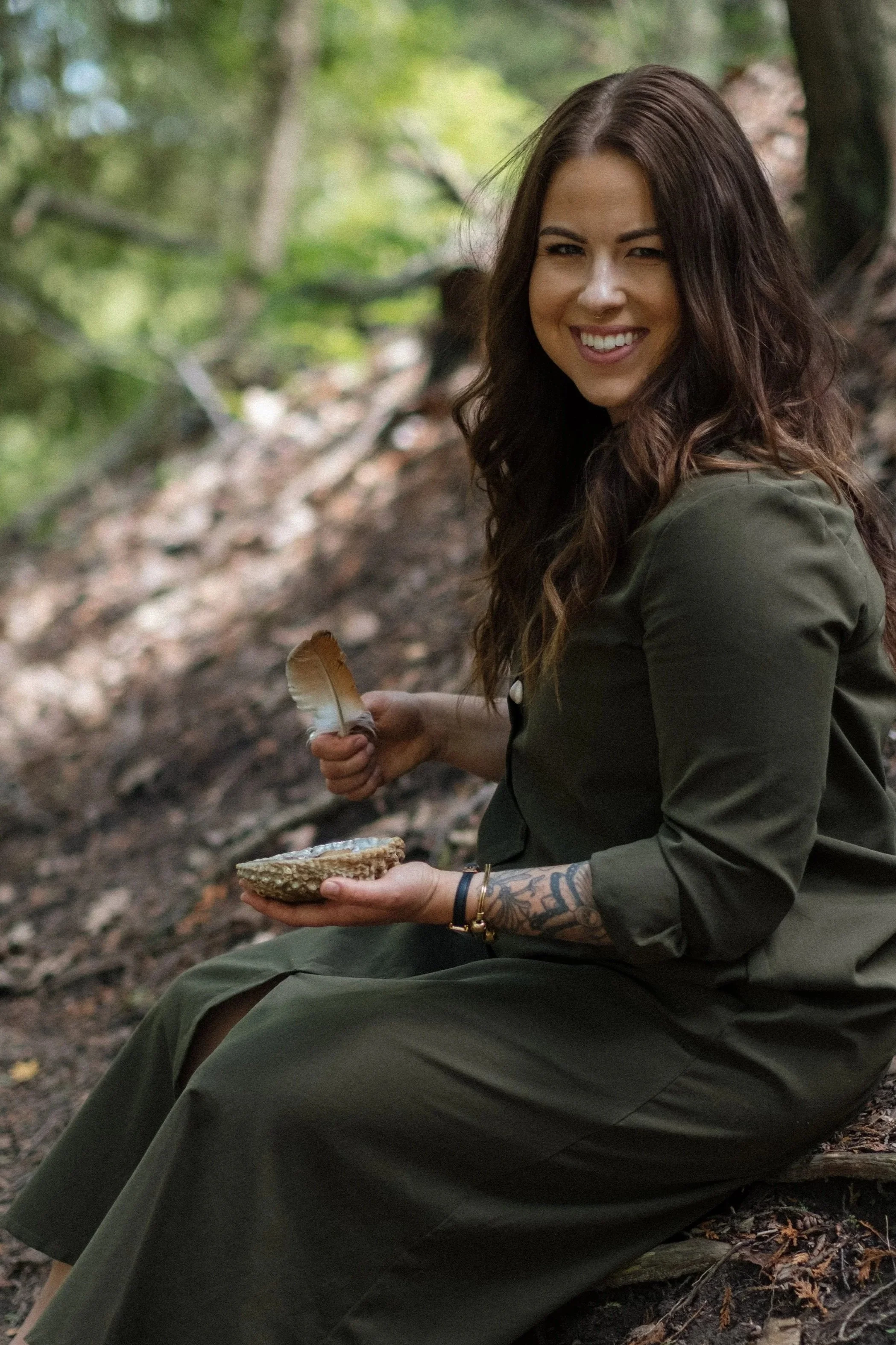 A woman with long brown hair and tattoos on her right arm sitting outdoors in a forest, smiling, holding a feather in one hand and a bowl with a flat object in the other.