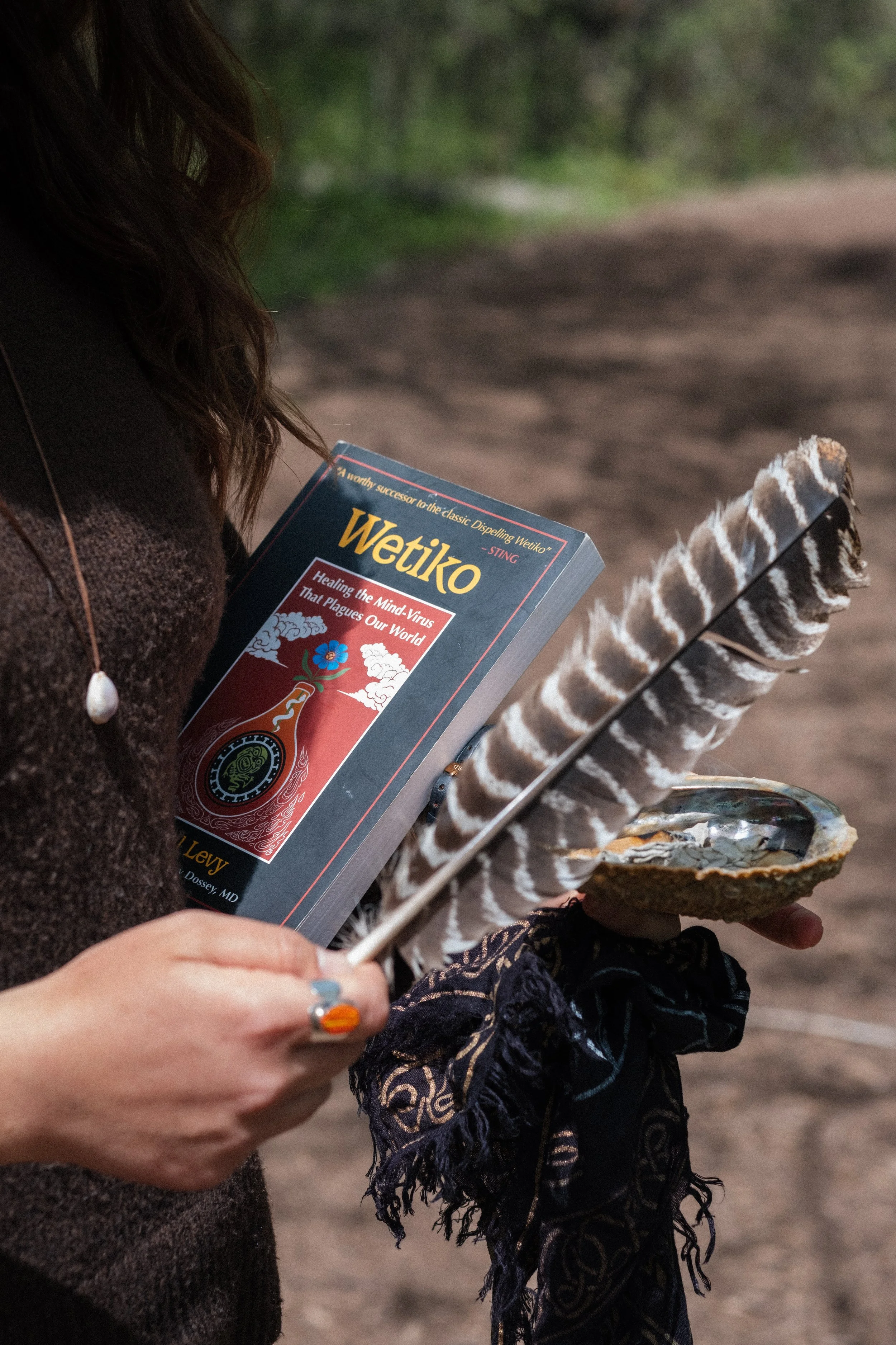 Person holding a book titled 'Wetiko' and a large feather, with an open shell on their hand, outdoors.