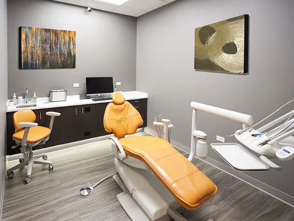 Dentist examination room with an orange dental chair, a computer, a yellow chair, and abstract artwork on the walls.
