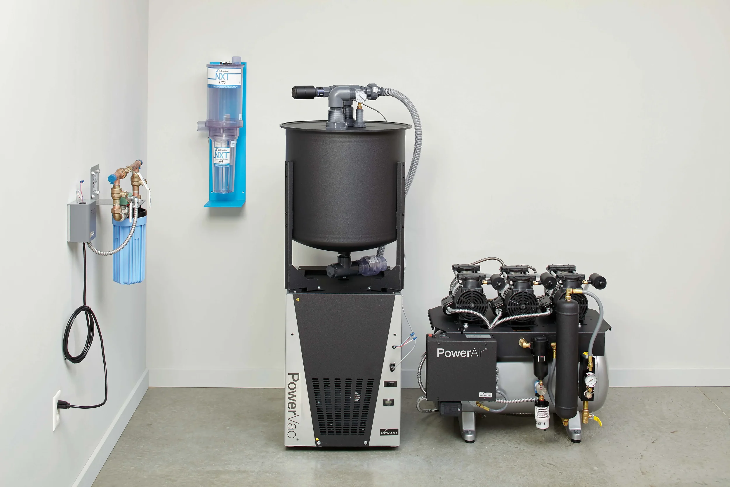 Medical or laboratory equipment with a black tank, blue filter system, and compressor in a room with white walls and gray flooring.