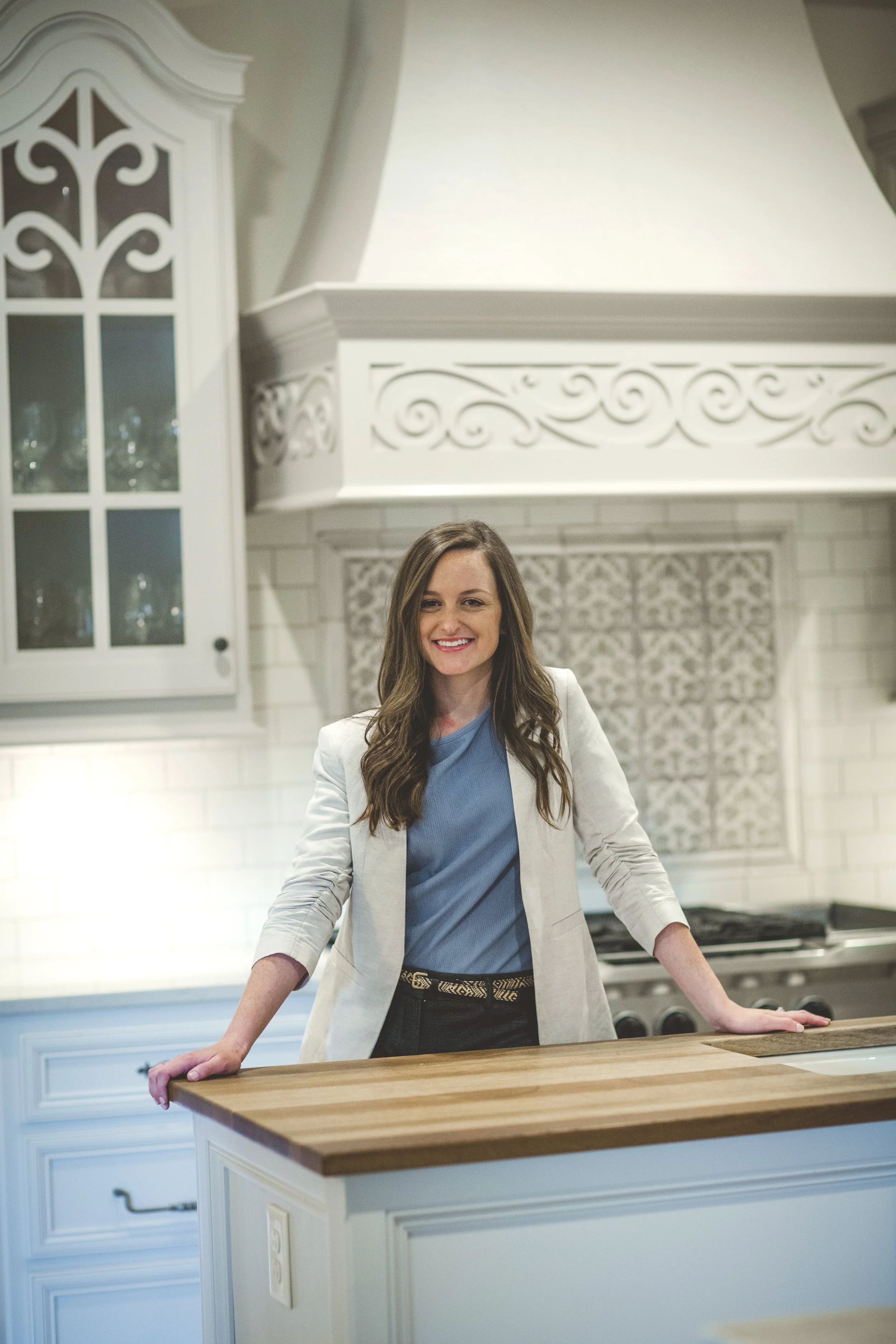 A smiling woman with long brown hair, wearing a blue top and white blazer, stands at a kitchen island in a bright, white kitchen with decorative cabinetry and a large oven range.