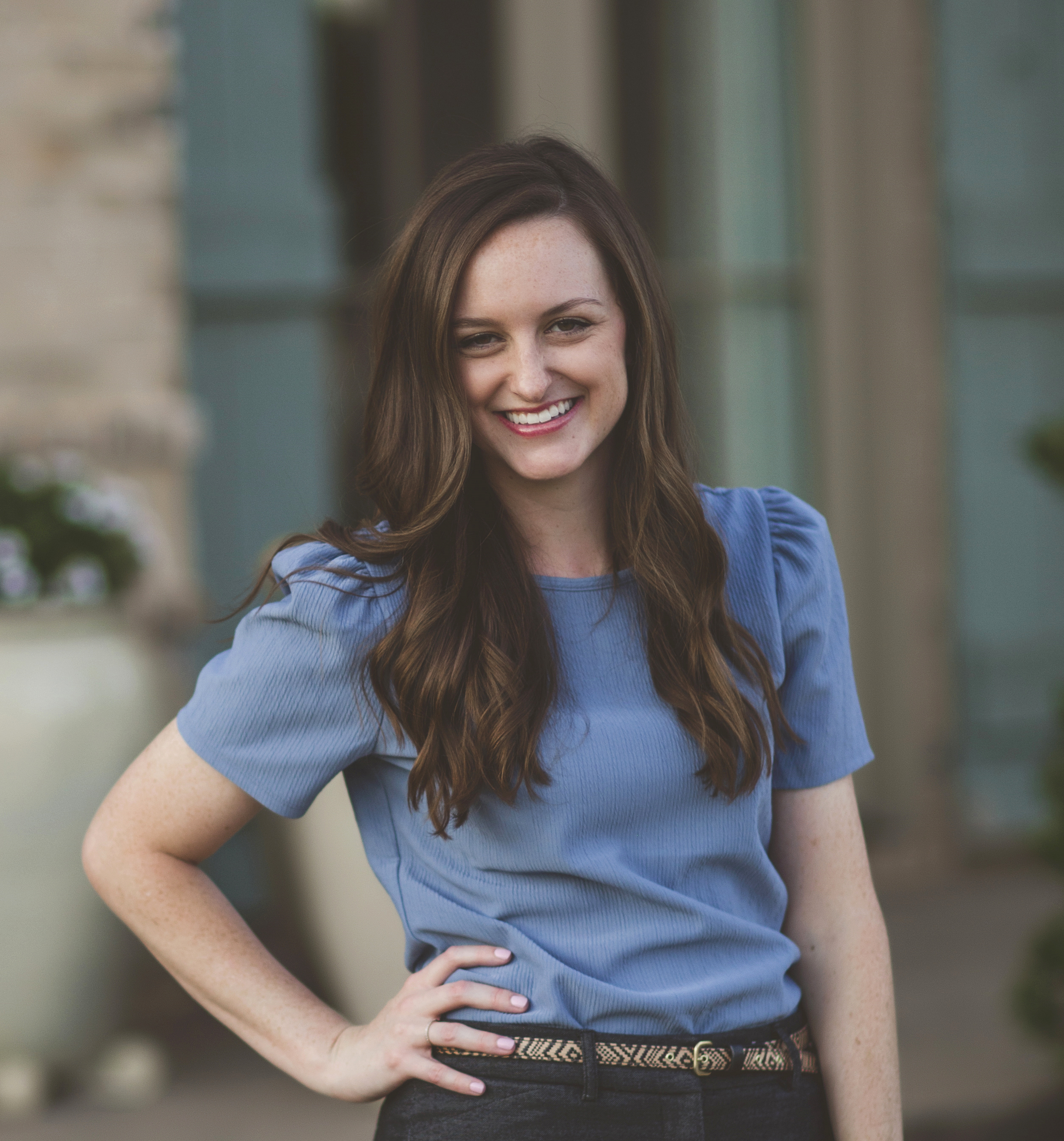 A woman with long brown hair smiling, wearing a blue short-sleeve top and dark pants standing outdoors.