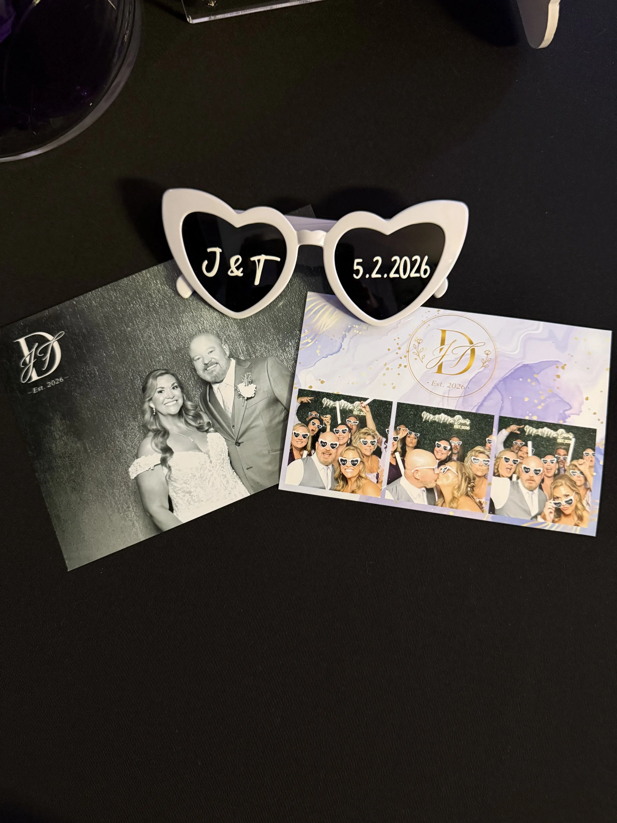 A pair of white, heart-shaped sunglasses with black lenses, displaying the initials on the left lens and the date on the right lens, surrounded by wedding-themed photos and decorations. Aubrey, TX wedding.