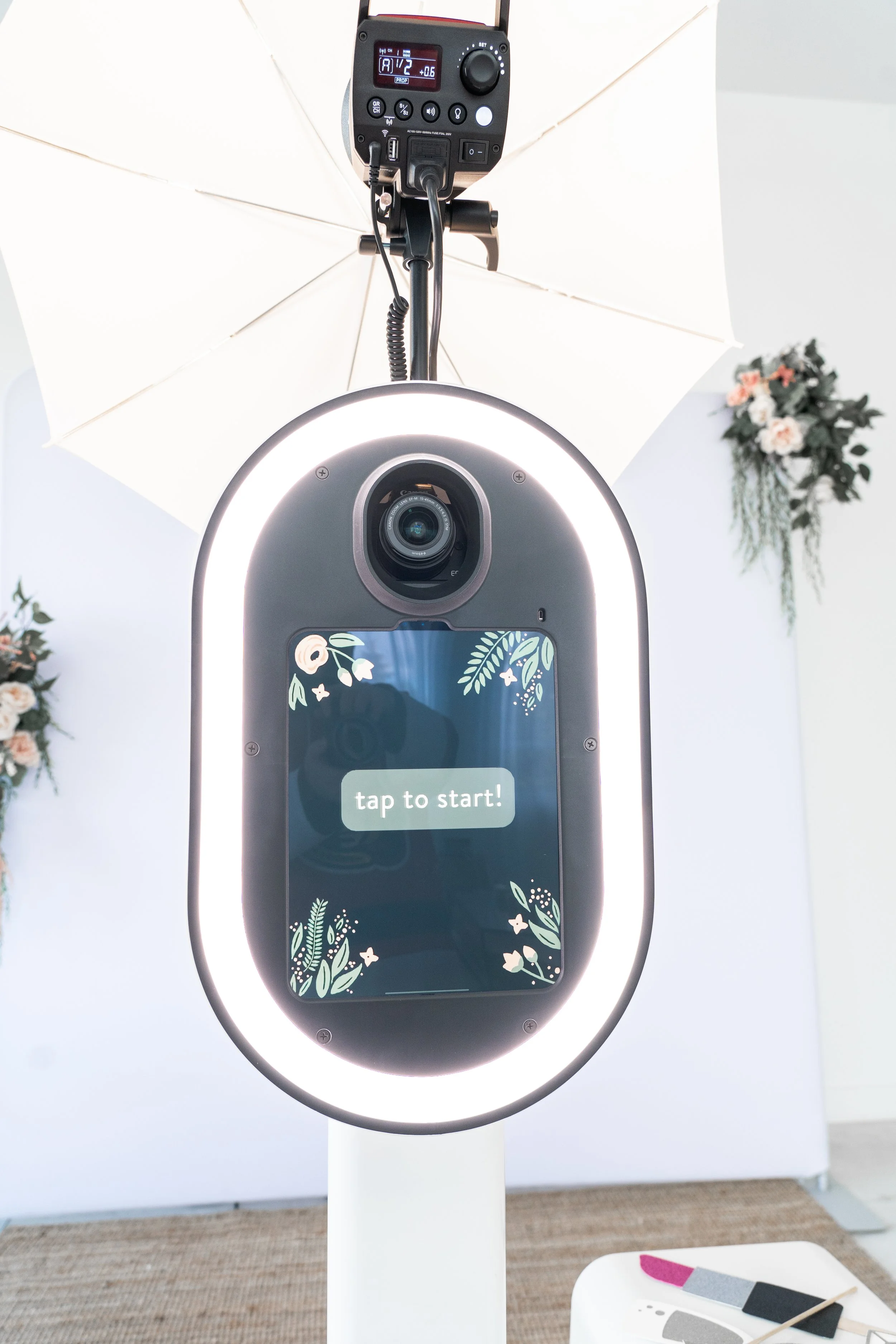 Photo of a modern photo booth with a screen displaying 'tap to start!' surrounded by floral designs, under a white umbrella with lighting equipment above, in a bright room decorated with flowers on the walls.