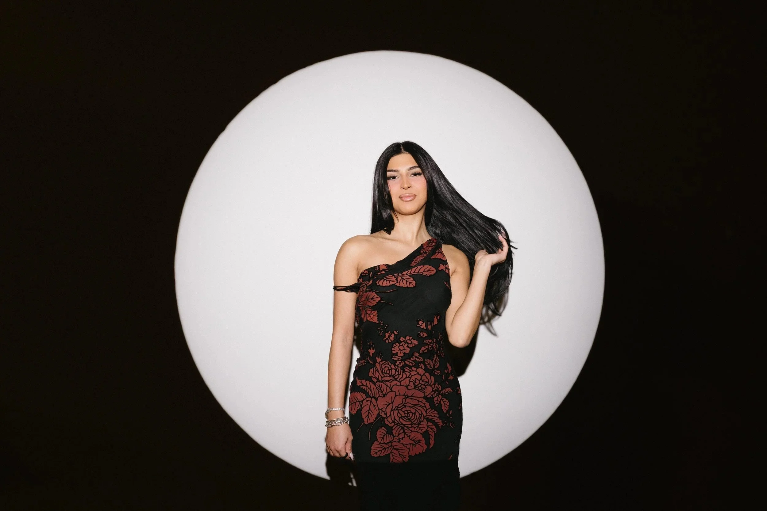 A woman with long black hair wearing a black dress with red floral pattern, standing in front of a large white circular backdrop with a black background.