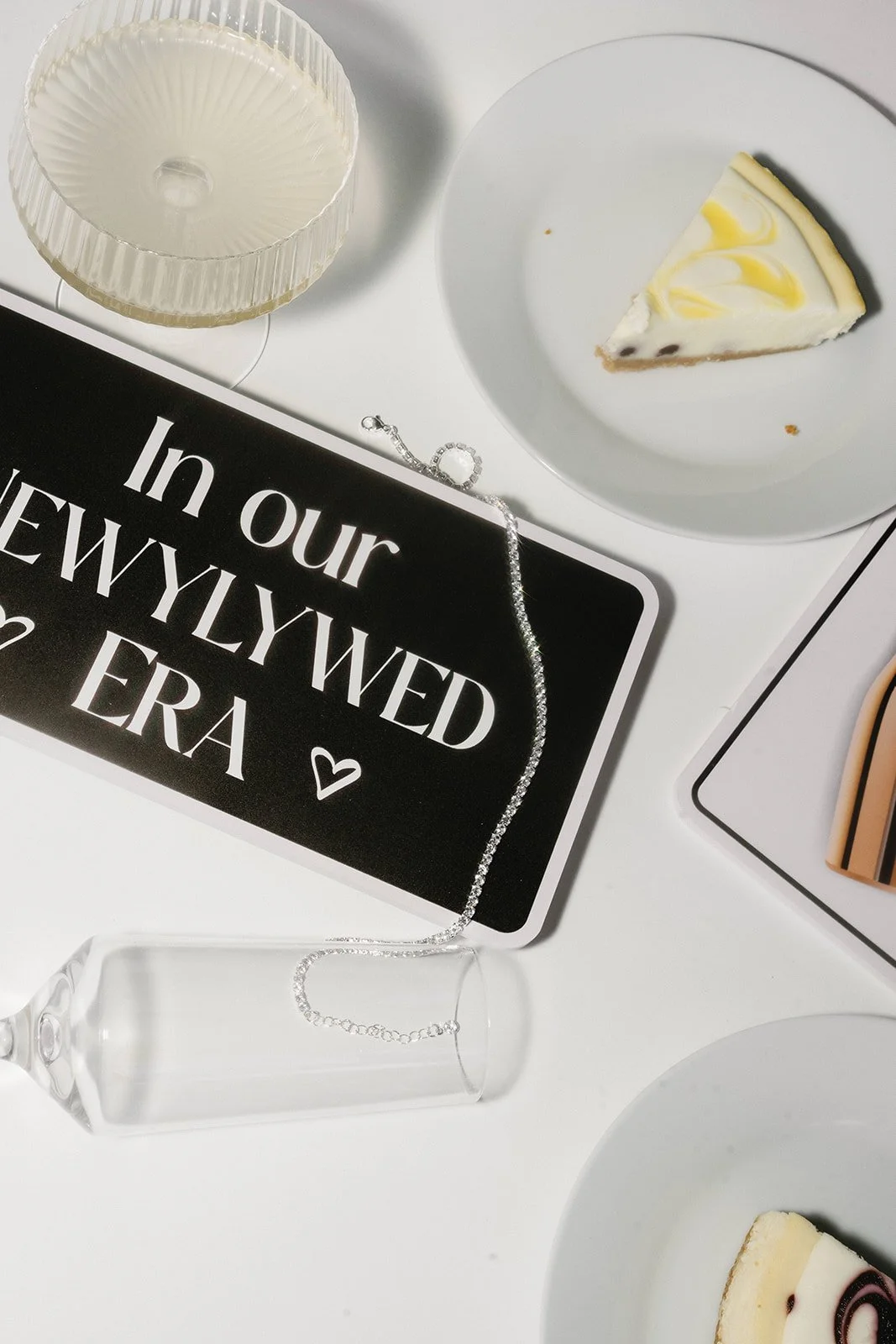 A celebration setup with a black sign that reads 'In our newlywed era' holding a silver necklace, a white cupcake wrapper, two slices of cheesecake on white plates, and a champagne flute lying on its side.