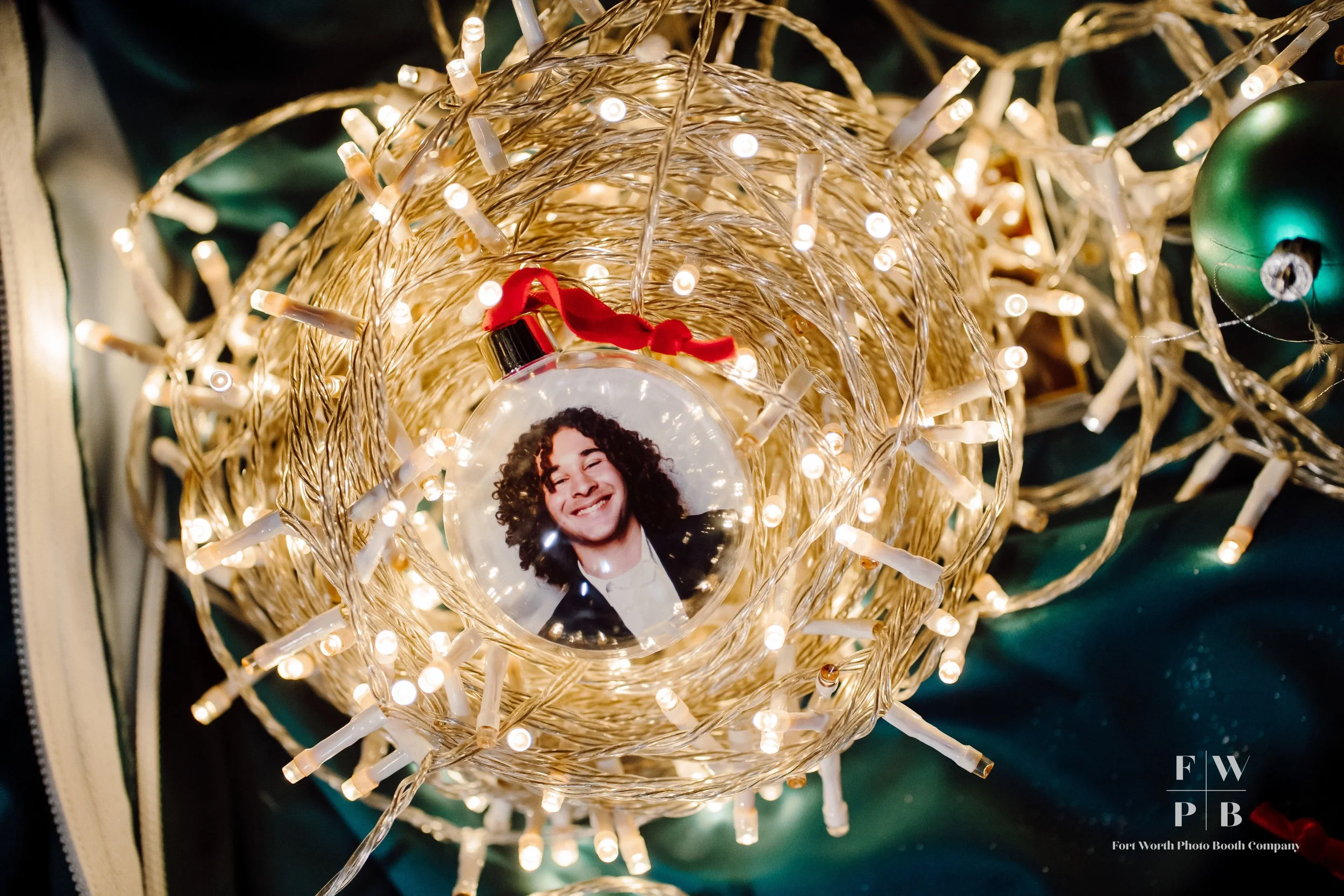 Close-up of a decorative string of white Christmas lights intertwined with a photo ornament of a smiling woman with curly hair, with a red bow on the ornament, and a green ornament in the background.