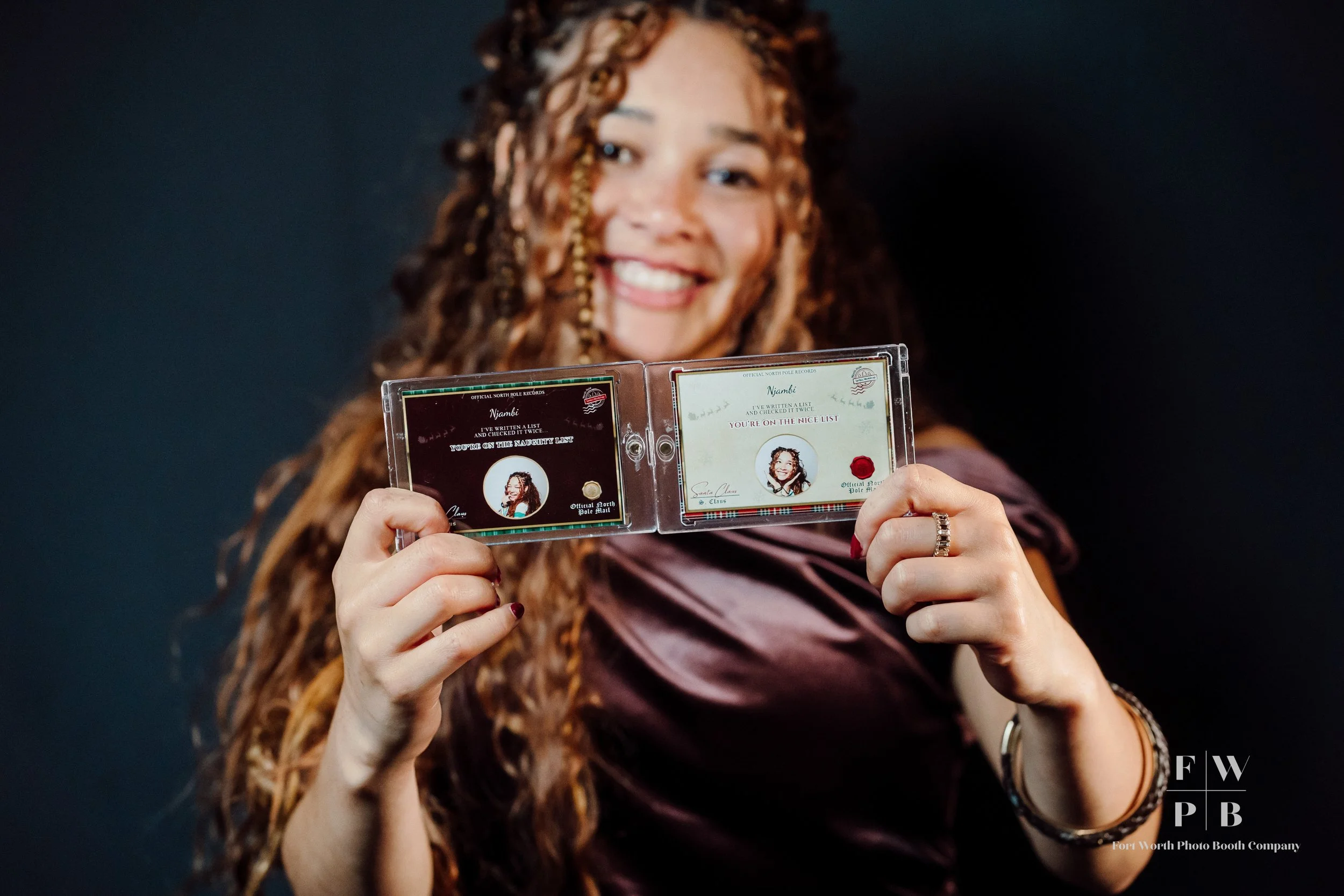 A woman with long, curly hair smiling and holding open a passport-sized photo ID, showcasing it to the camera against a dark background.