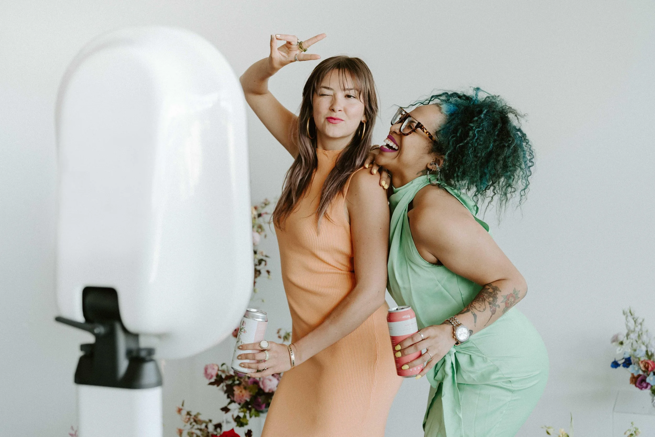 Two women taking a selfie, one with brown hair in a peach dress, making a peace sign and winking, the other with curly teal hair in a green dress, smiling and leaning into her friend, holding canned drinks, surrounded by flowers.