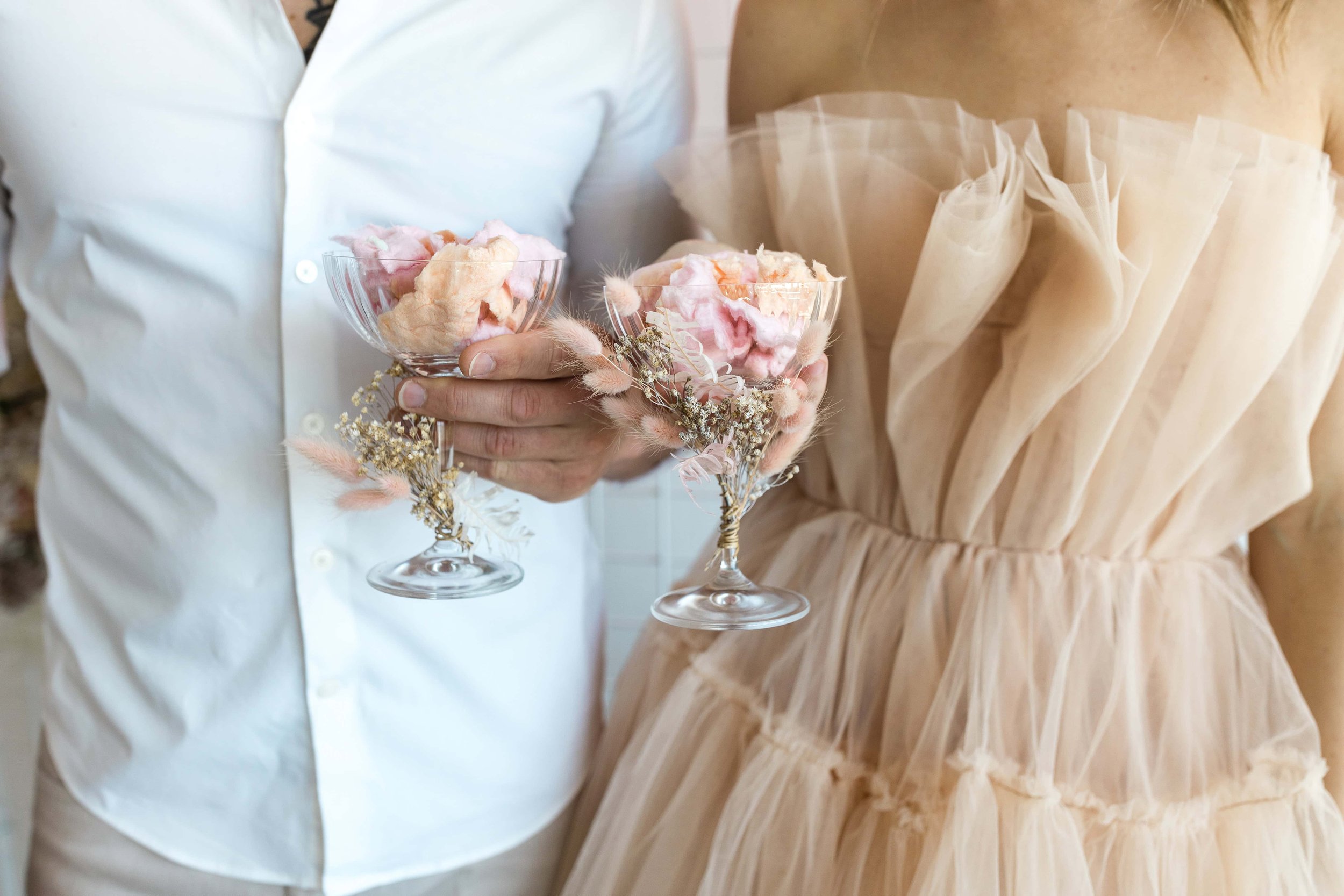 People holding glass bowls filled with pink cotton candy, with one person wearing a light-colored dress and the other in a white shirt.