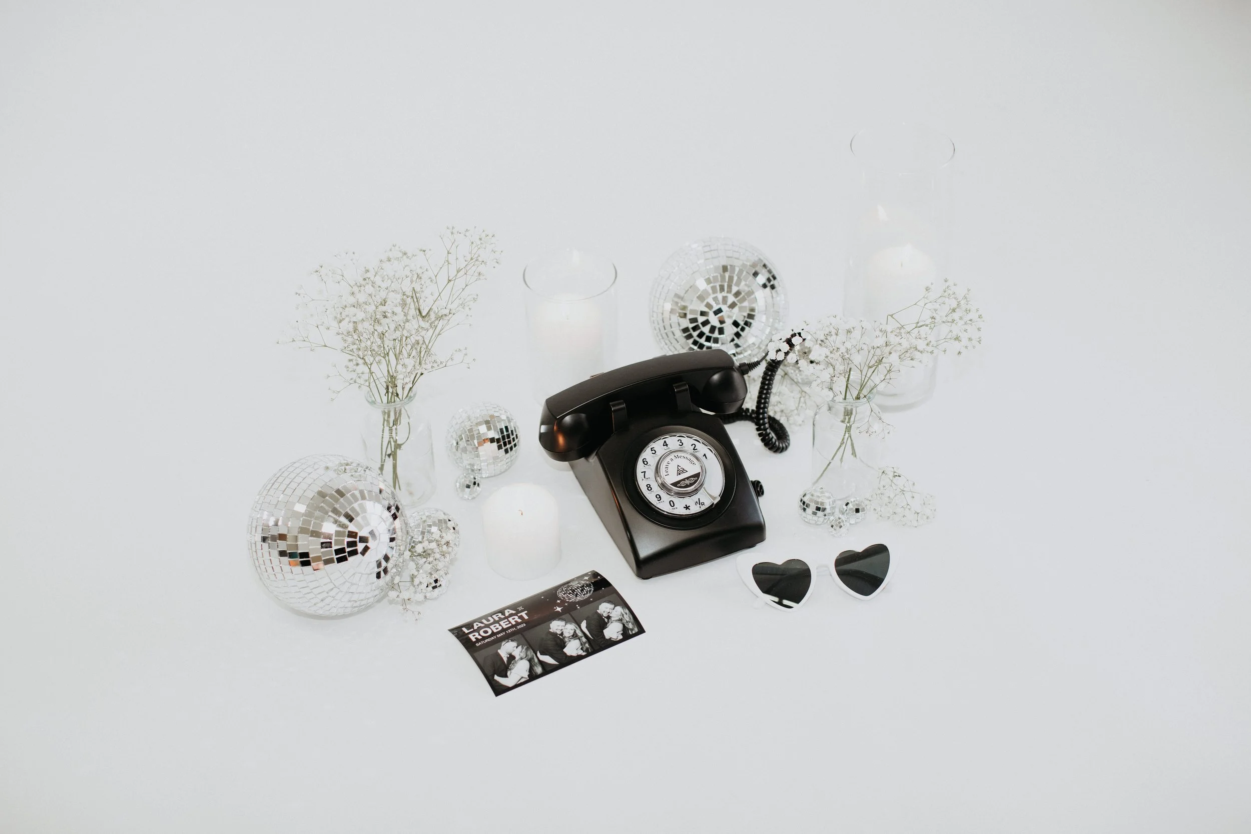 Vintage black rotary telephone surrounded by small disco balls, candles, flower vases with dried baby's breath, heart-shaped sunglasses, and a photo card with black and white images.