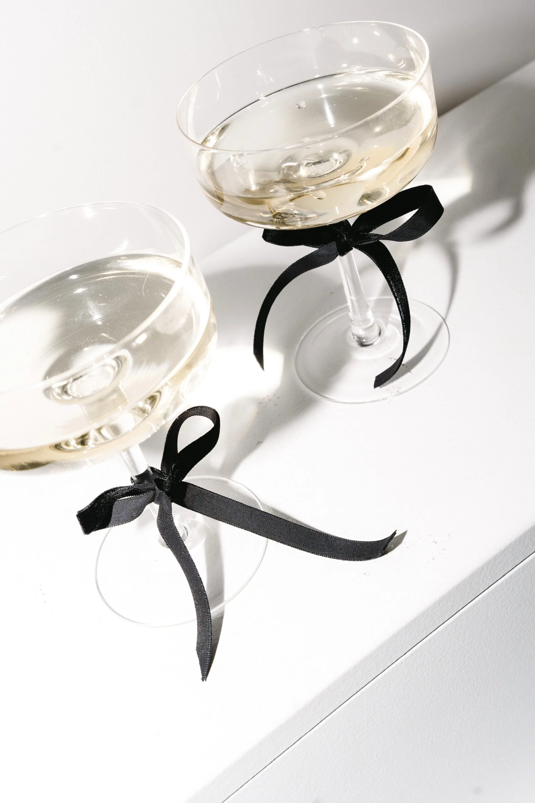 Two champagne glasses with black ribbons tied around the stems, filled with white wine, on a white surface.