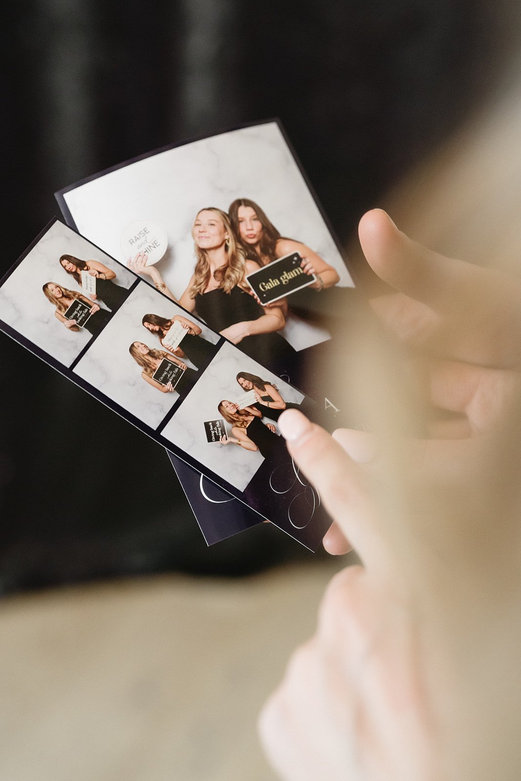 Photograph of a person holding a portfolio with multiple photos of two women at a photo booth. The photos include the women smiling, holding signs, and interacting, against a light gray background.
