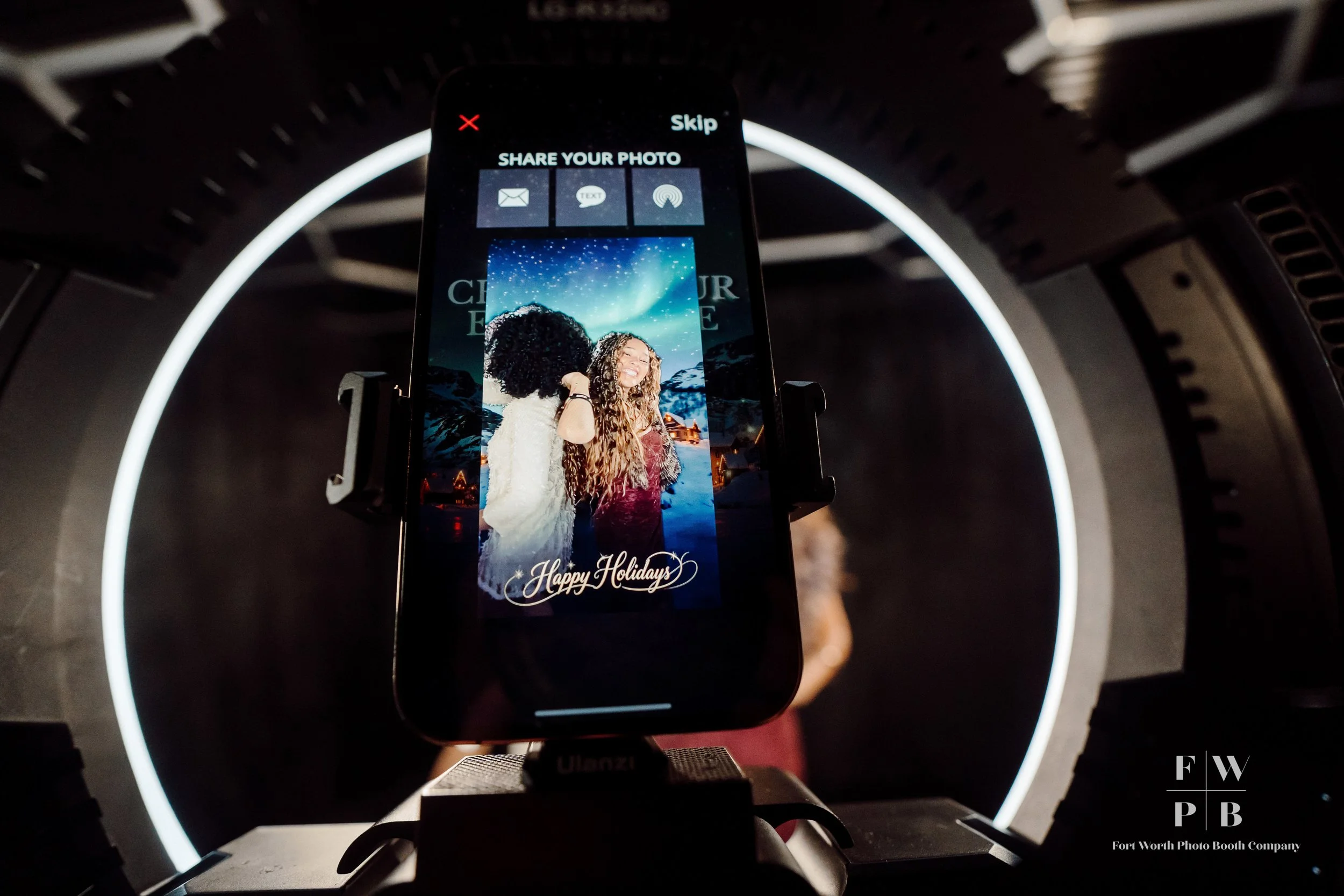 A smartphone displaying a holiday photo of two women smiling and hugging, with a starry sky and northern lights in the background, inside a light ring setup for photography.