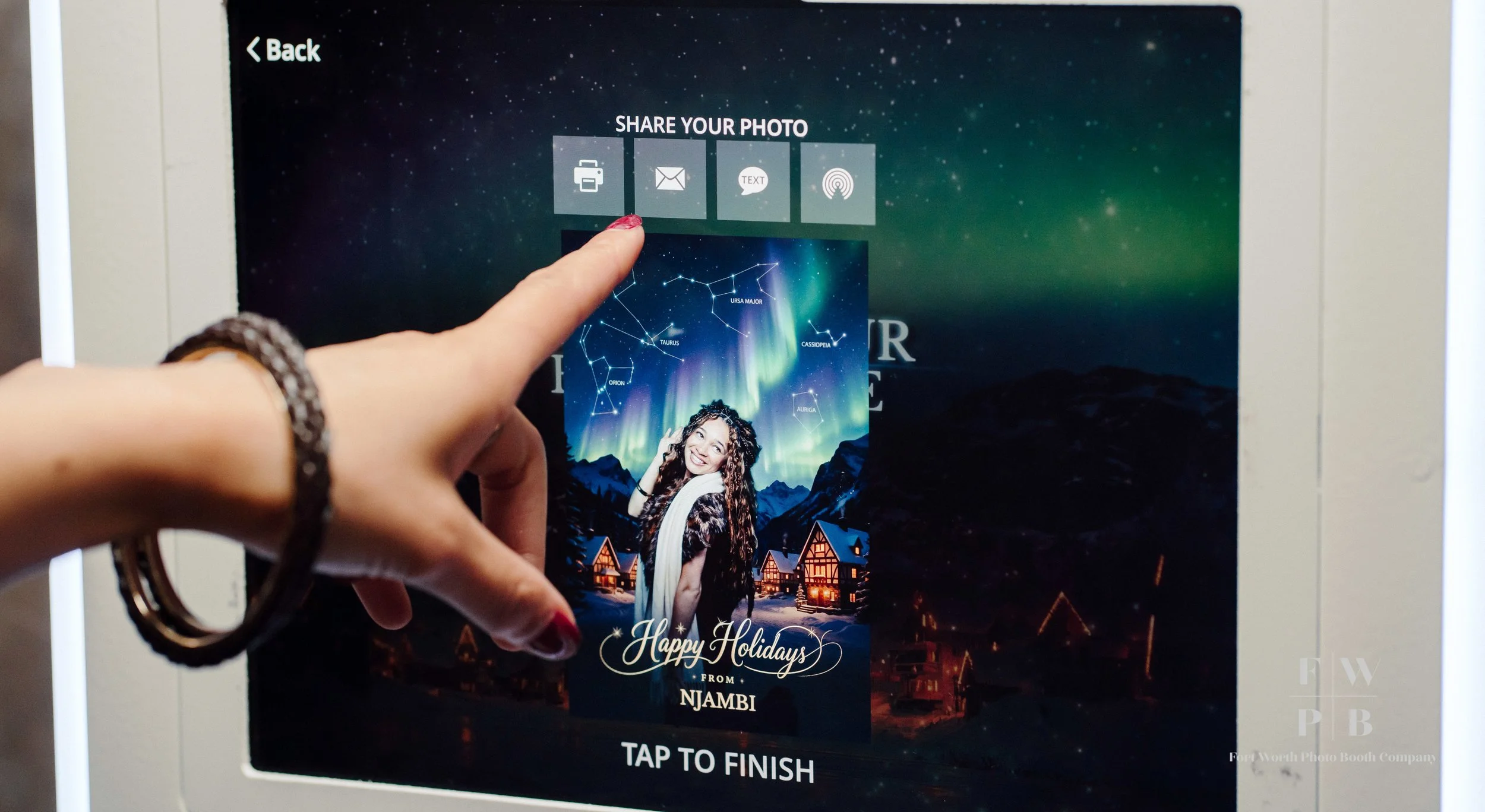 Person pressing a photo booth touchscreen with a holiday card featuring a girl in front of a mountain and northern lights, with a cityscape at night, and options to share via printer, email, text, or social media.
