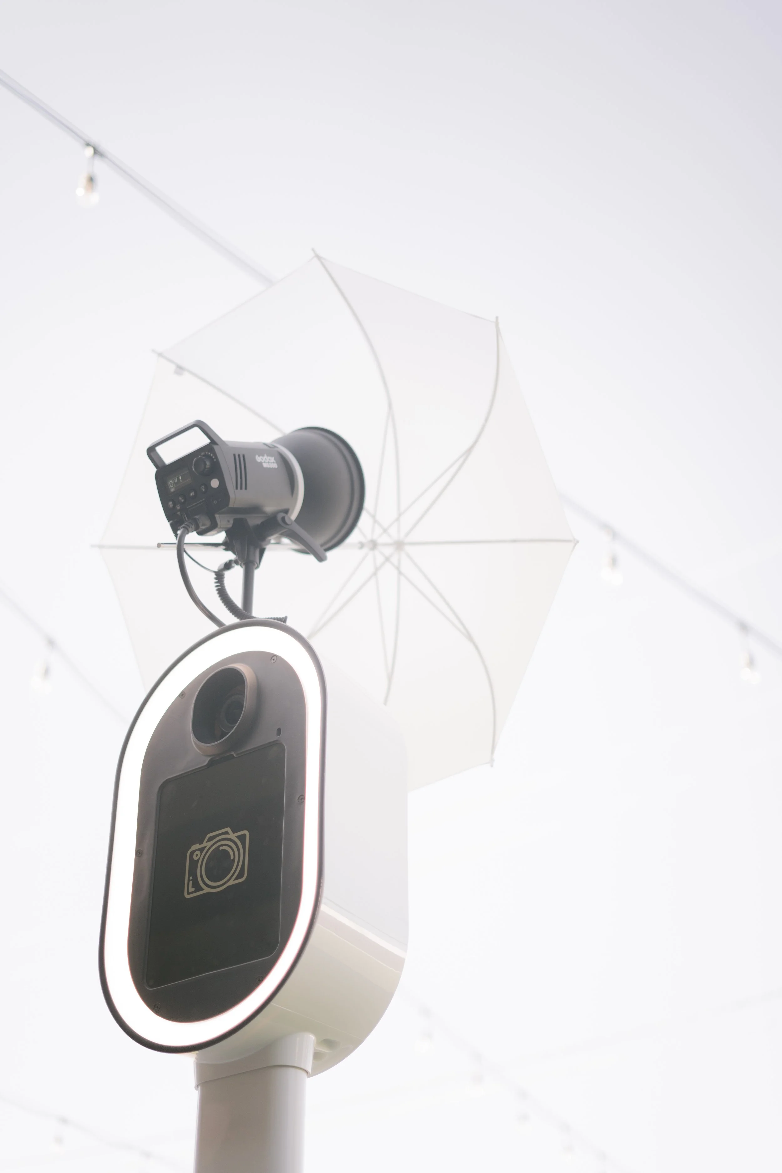 A photo of a photo booth with a large ring light, camera, and an umbrella for lighting, all mounted on a white stand.