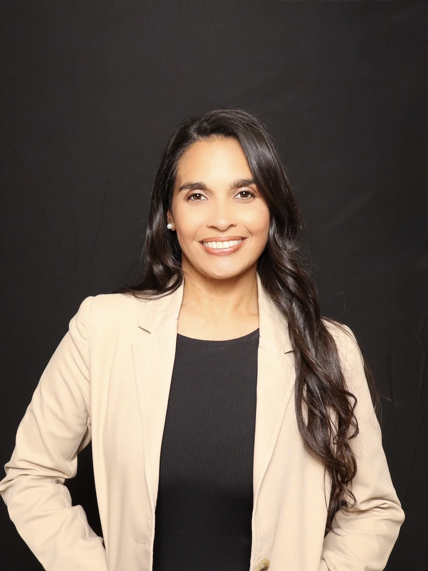A woman with long dark hair, wearing a beige blazer and a black top, smiling and standing against a black background.
