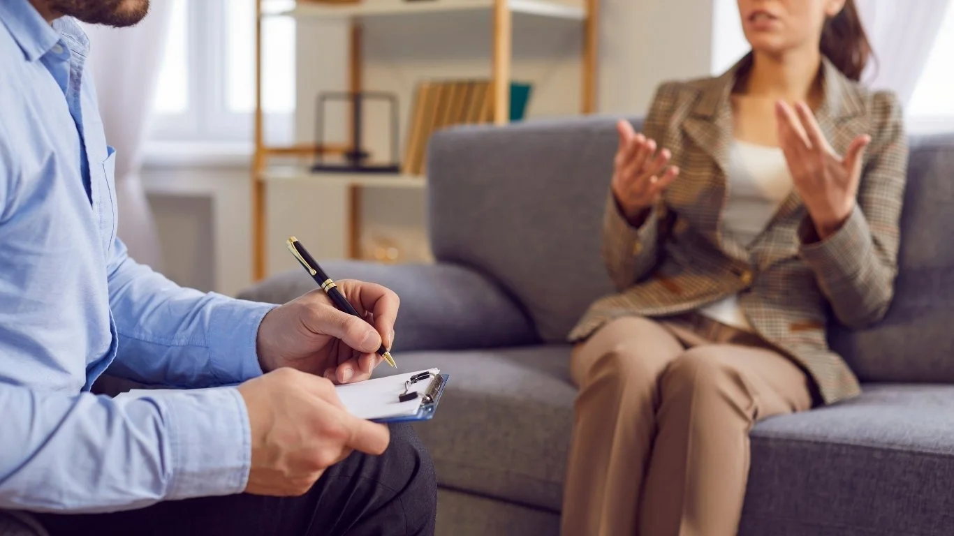 A woman in a plaid blazer and beige pants is talking to a therapist, who is taking notes on a clipboard during a therapy session in a bright office.