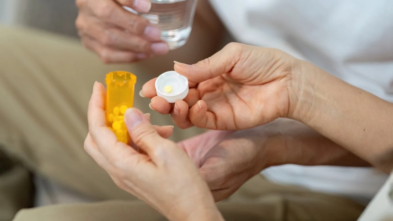 Hands holding a pill bottle and an open white case with yellow pills, with another hand holding a glass of water