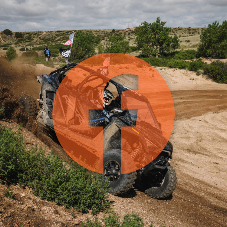 UTV driving on a dirt trail in a rural area with green shrubs and trees, under a cloudy sky, with onlookers and American flags in the background.