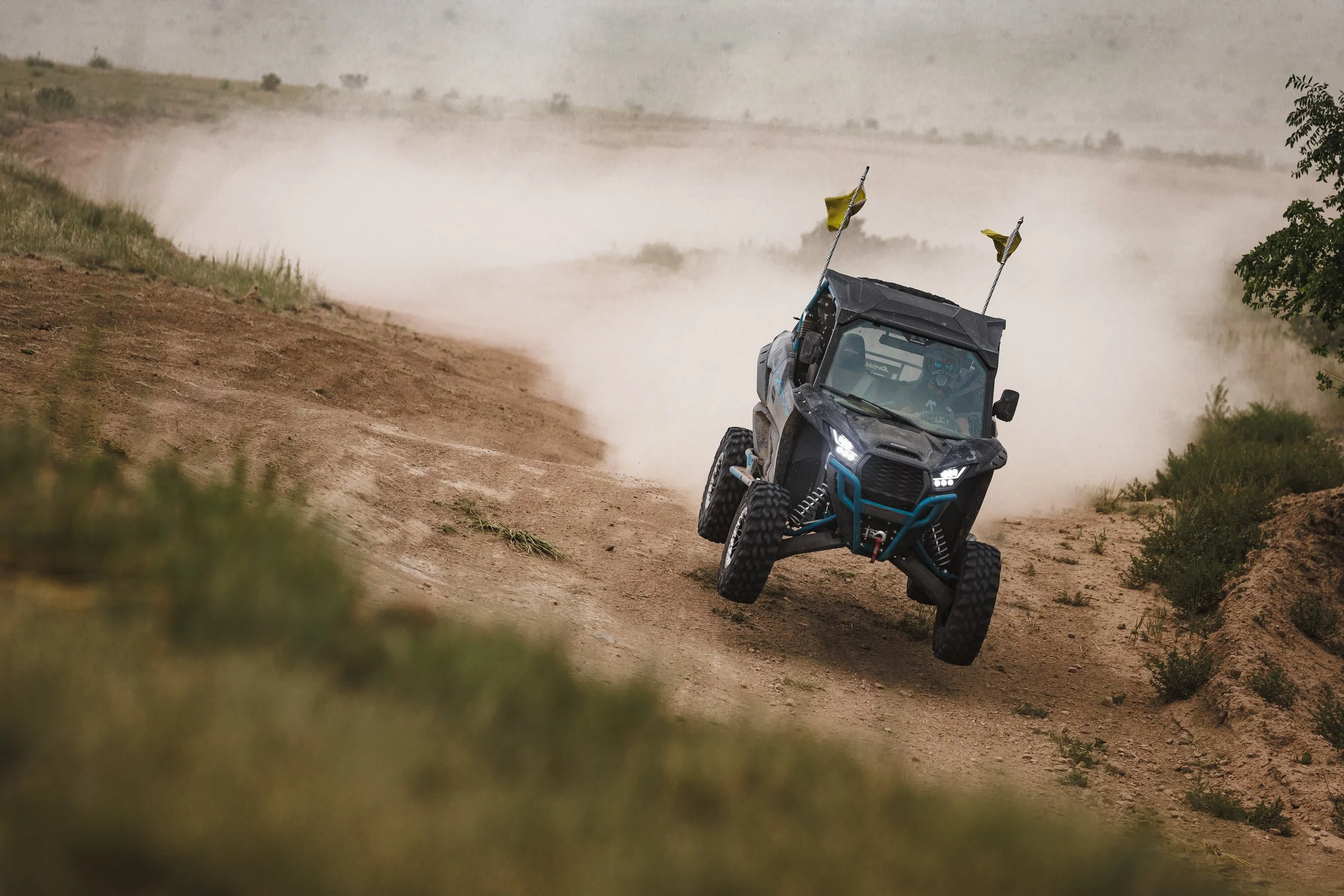 An off-road utility vehicle driving fast on a dirt trail, kicking up dust in a rural, grassy landscape.