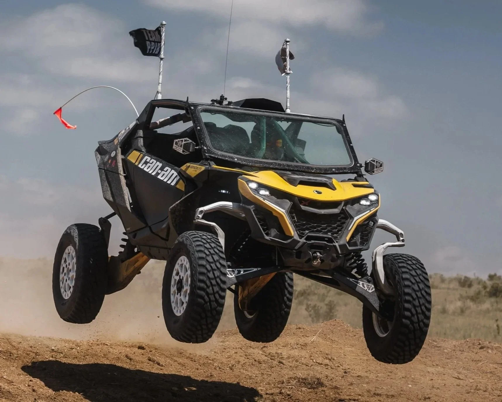 An off-road vehicle airborne over dirt terrain, with two flags attached to the vehicle, one with the text 'Team PP' and another with a cap on it.
