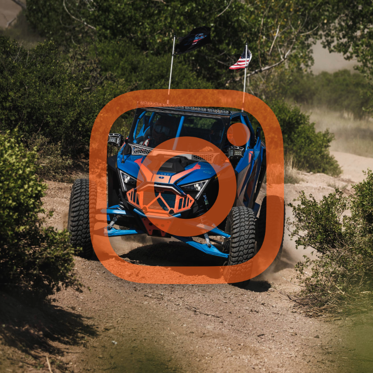 Blue off-road dune buggy with flags driving on dirt trail through desert plants.