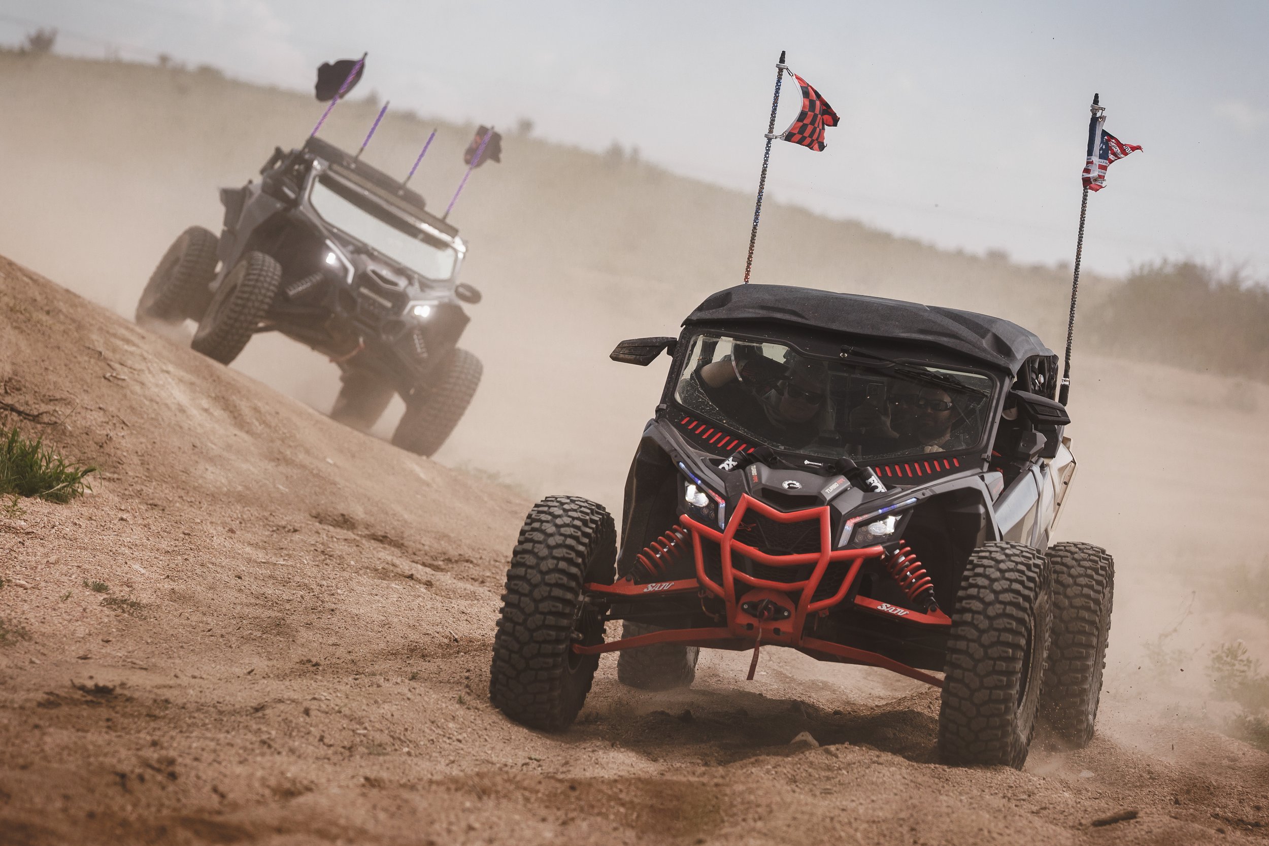Two off-road utility vehicles driving on a dirt trail in a desert landscape, kicking up dust, with flags mounted on their roofs.