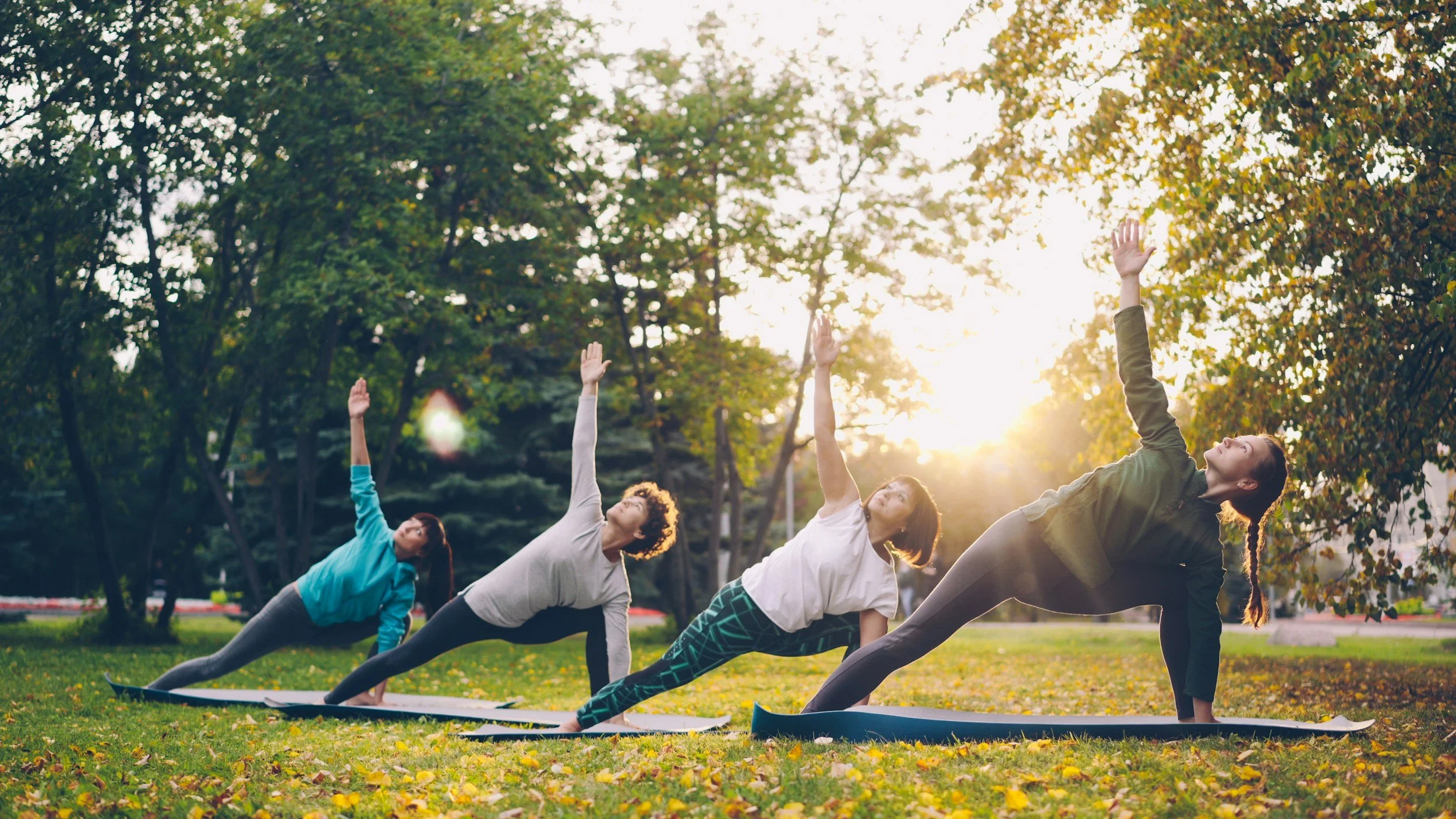 Four women practicing yoga outdoors on mats in a park during sunset, performing side plank poses with one arm raised, surrounded by trees and fallen leaves.