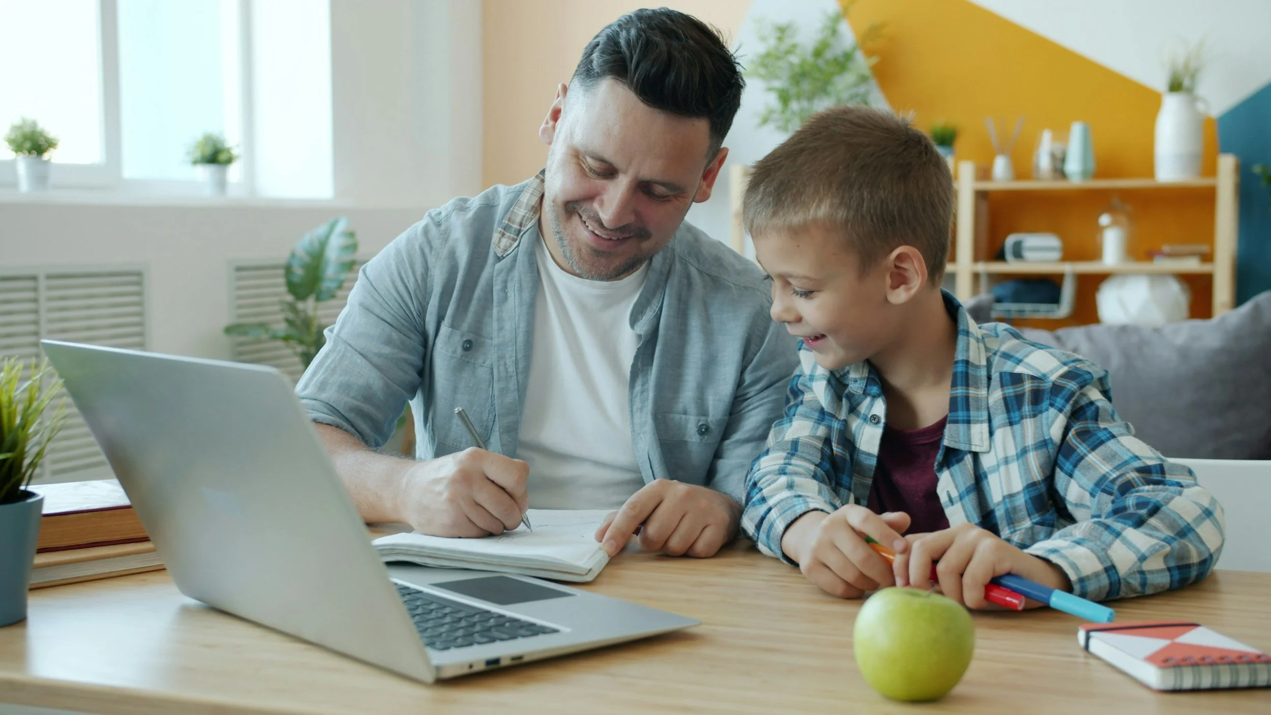 A man and boy smiling and looking at each other while sitting at a table with school supplies, a green apple, and a laptop in a bright room.