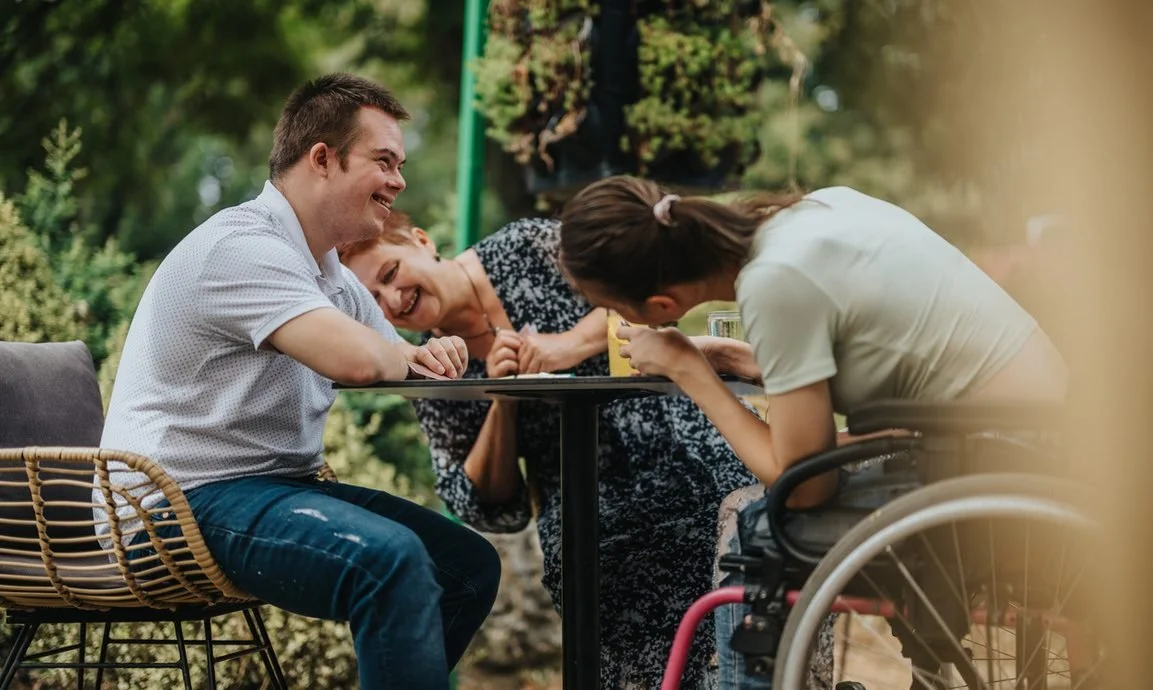 Three people laughing and smiling at a table outdoors, one woman in a wheelchair, two individuals leaning forward.