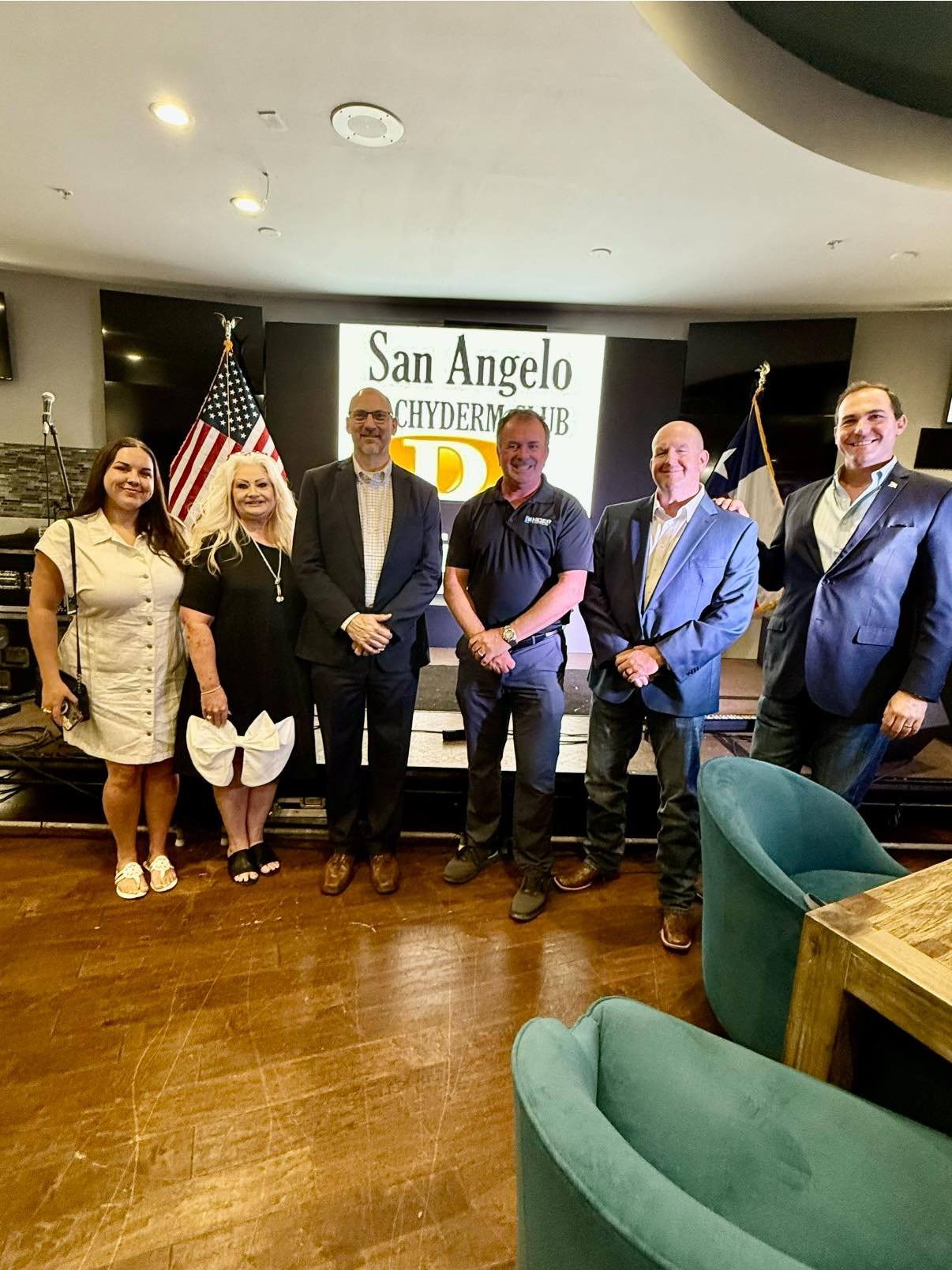 Group of six people standing on a stage in front of a sign that reads "San Angelo Chamber of Commerce". Two flags are behind them, one U.S. flag and one Texas flag.