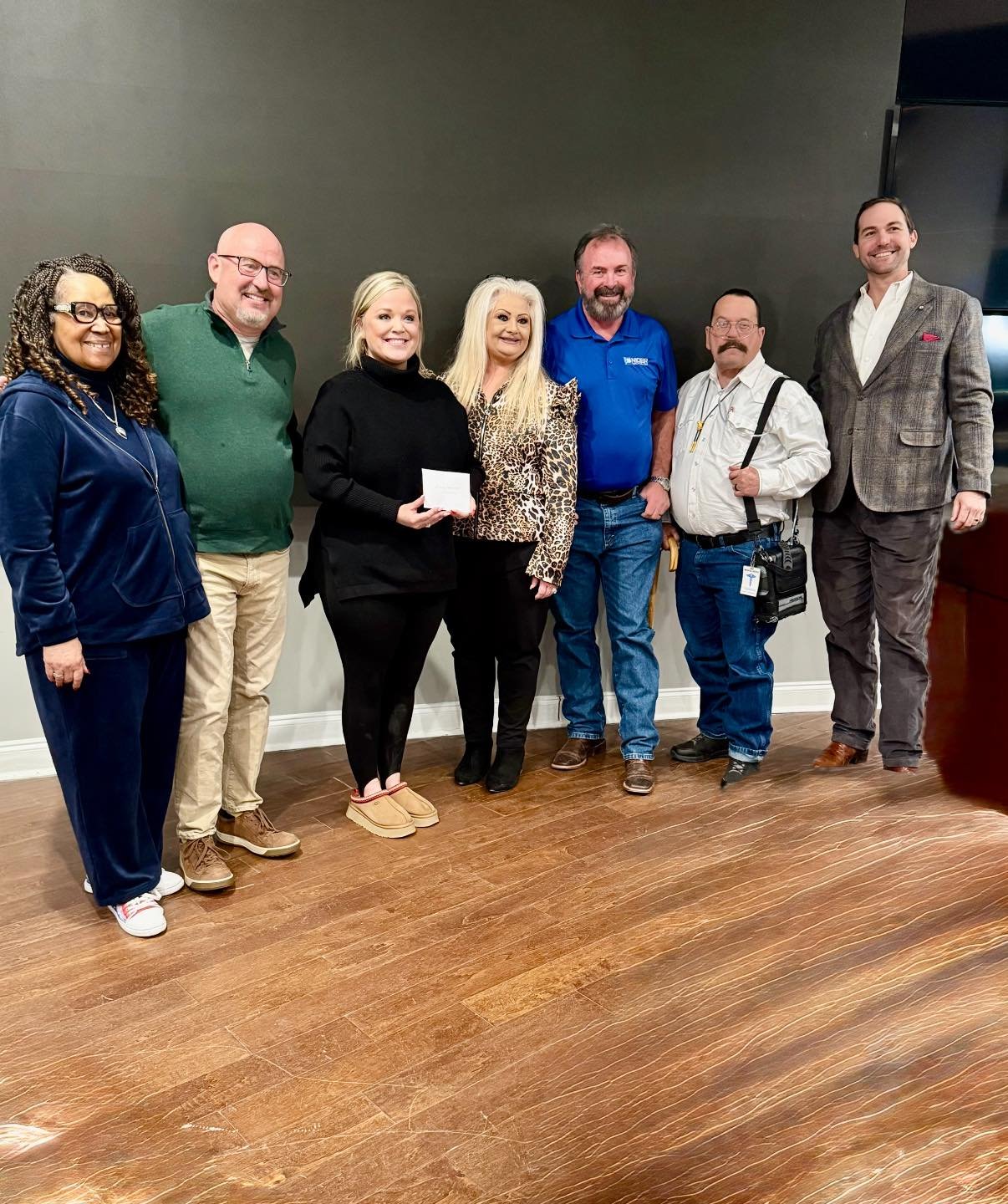 Group of seven people standing together indoors in front of a plain wall, smiling at the camera.