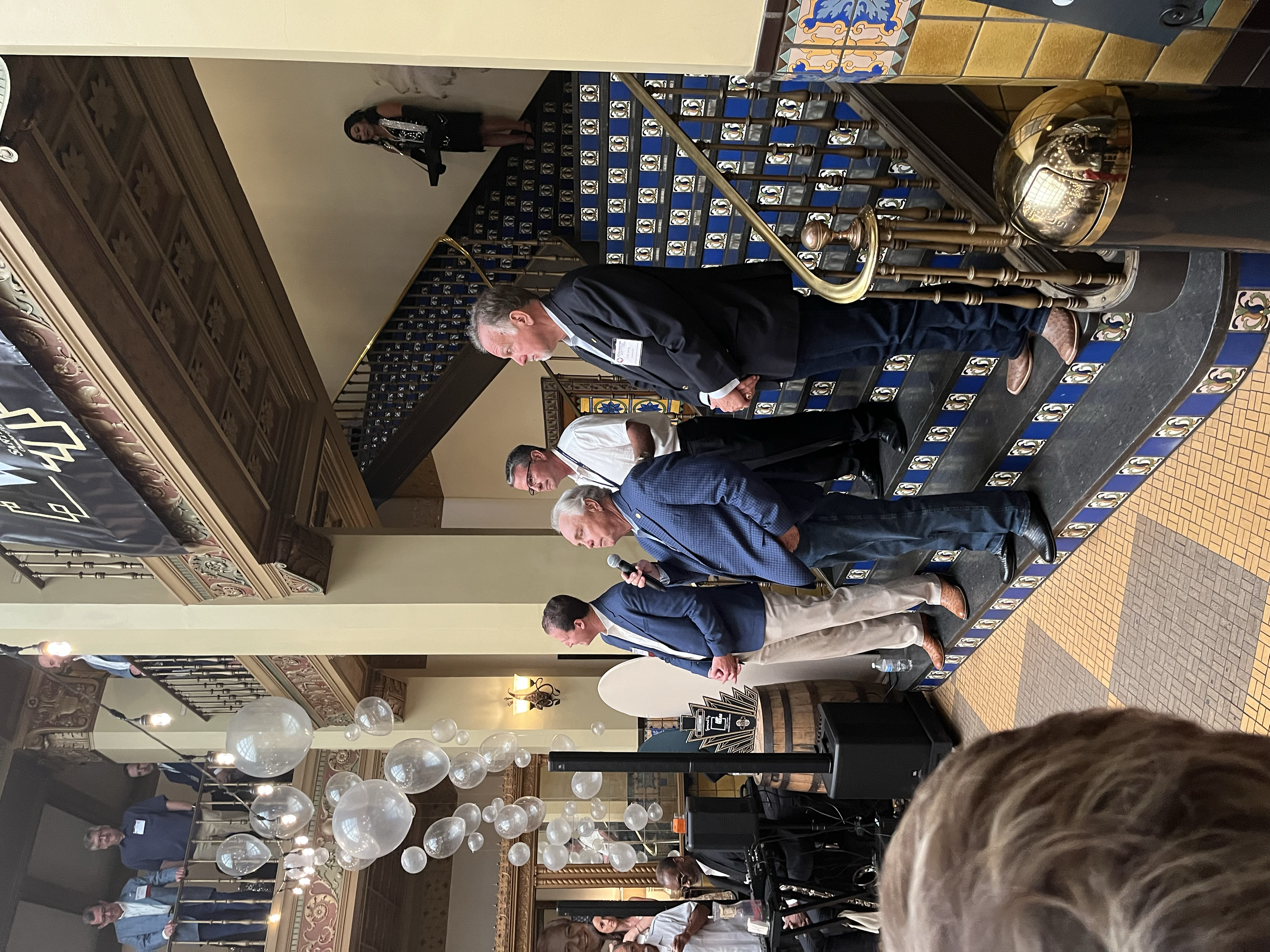 Four men in business attire standing on a decorated stage with a microphone and banners, engaged in conversation during an event in a hotel ballroom.