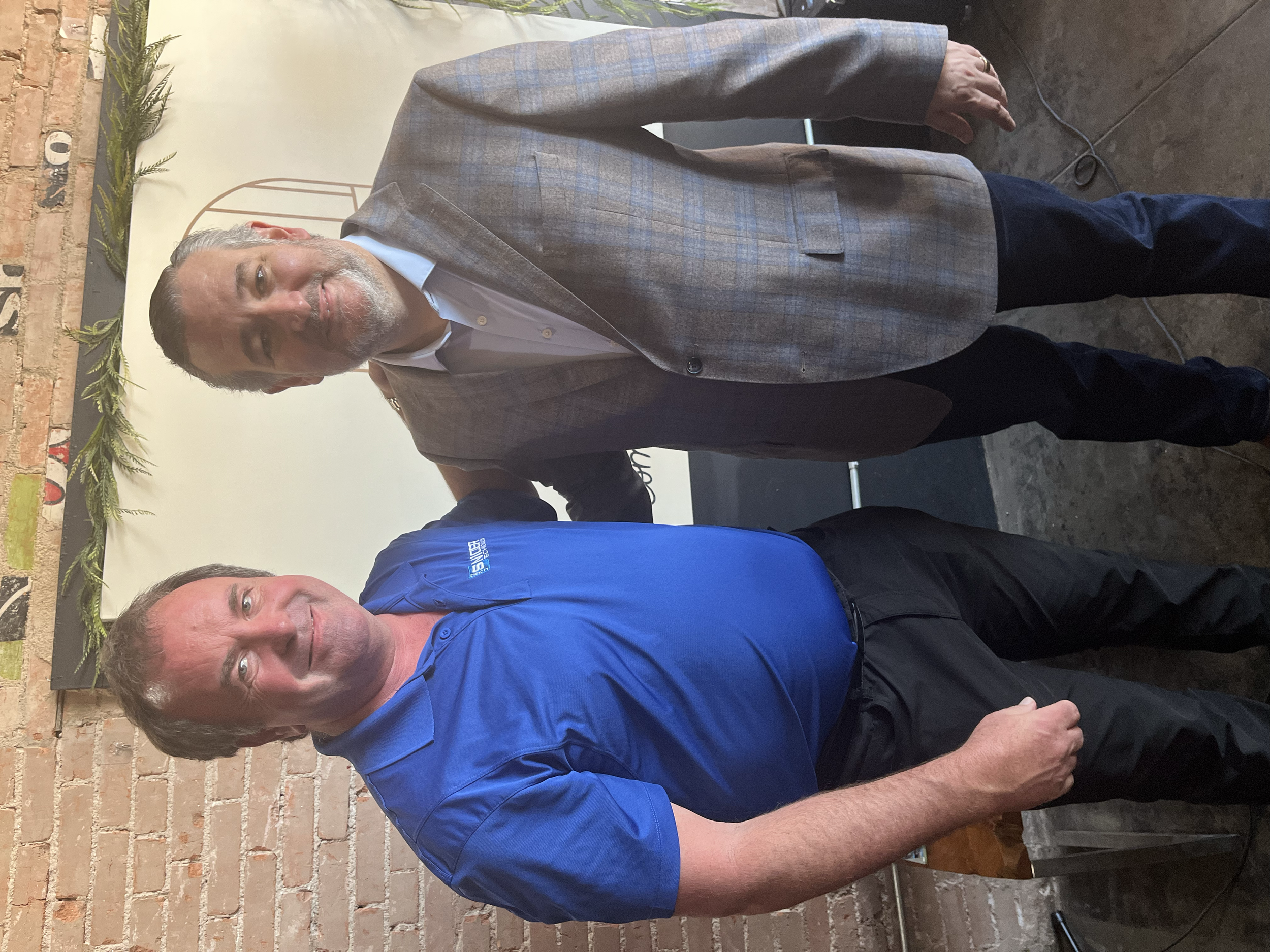 Two men standing indoors, one dressed in a blue shirt and black pants, and the other in a gray plaid blazer with dark pants. They are beside a table, with a brick wall and green plants in the background.