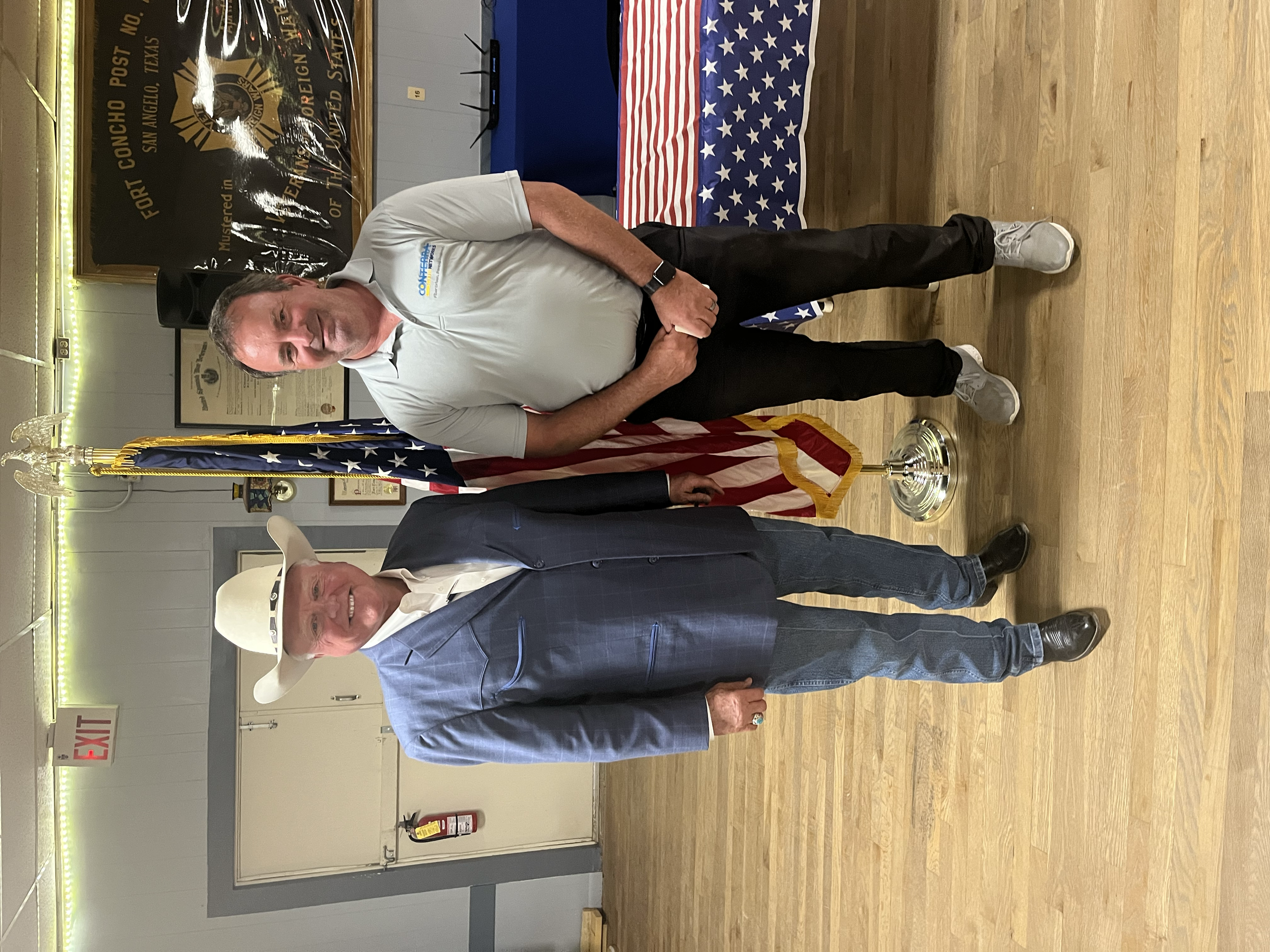 Two men standing indoors, one wearing a white cowboy hat, a blue plaid blazer, and jeans, and the other in a light gray shirt and black pants, with American flags behind them and a Concho County, Texas banner in the background.