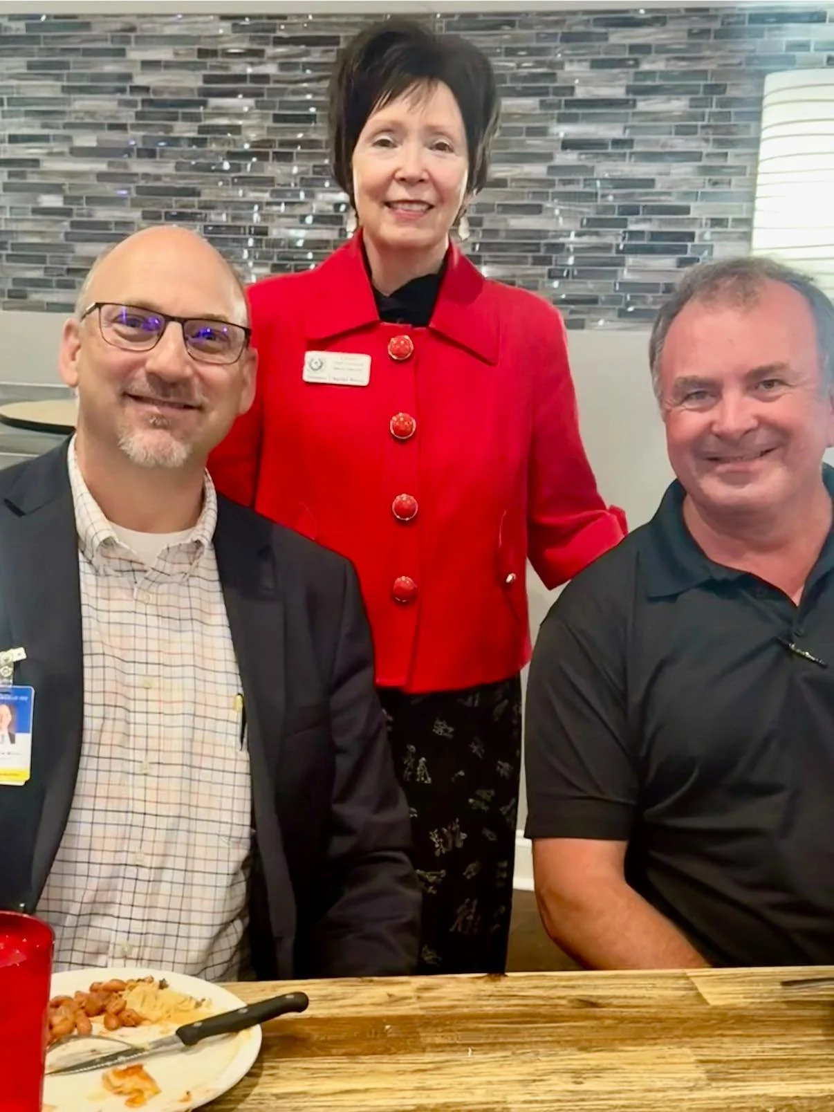 A woman in a red jacket standing behind two men sitting at a table in a restaurant.