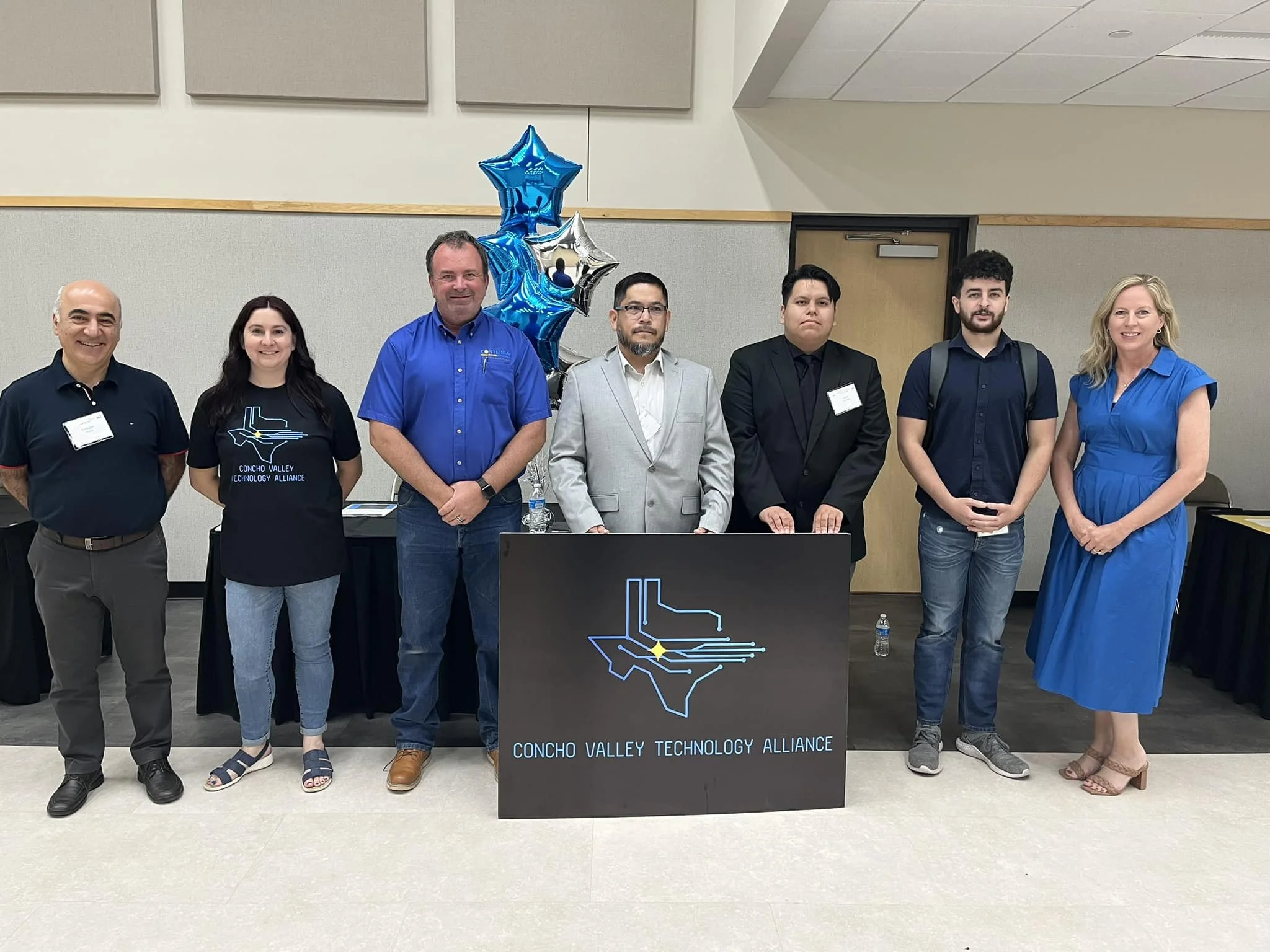 Group of seven people standing behind a desk with a sign that reads 'Concho Valley Technology Alliance.' There are blue and silver star-shaped balloons to the left of the group. The group includes men and women dressed in casual and formal attire, standing in a room with beige walls and a door in the background.